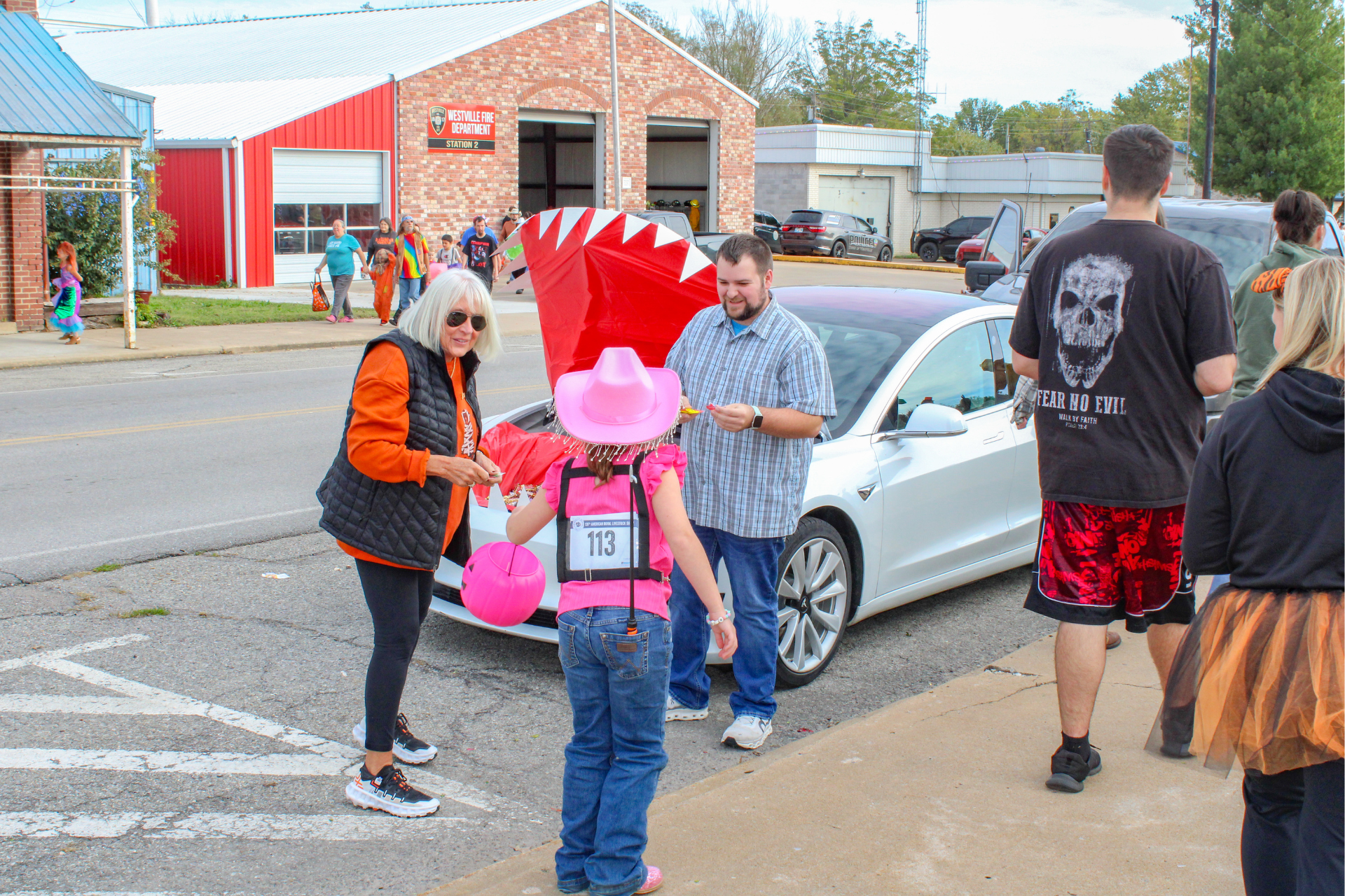 People in costumes gather near a white car on a street. A child in a pink hat and a person with a red headdress stand out.