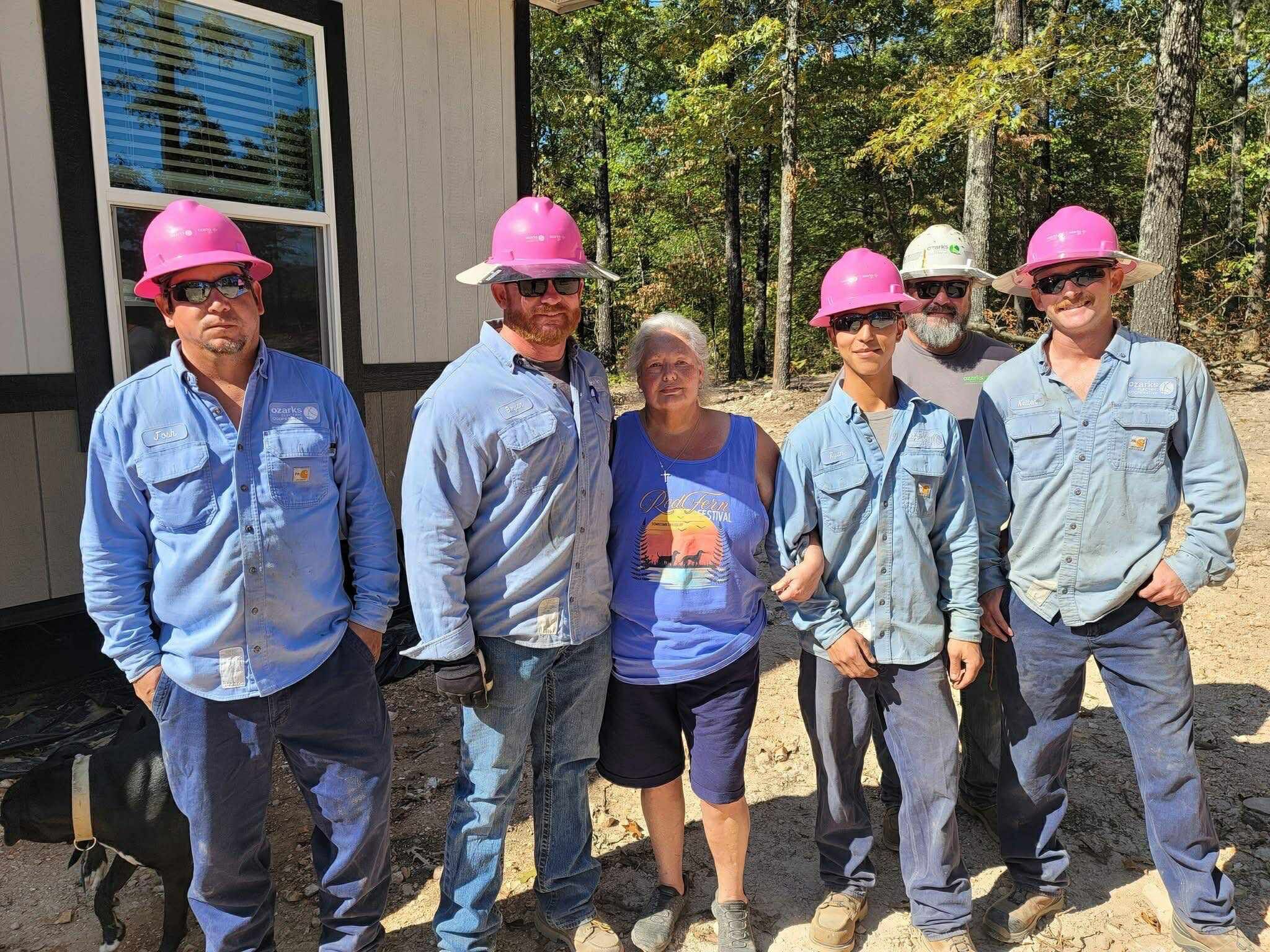 Five construction workers in pink helmets and a woman stand smiling in front of a building, surrounded by trees.