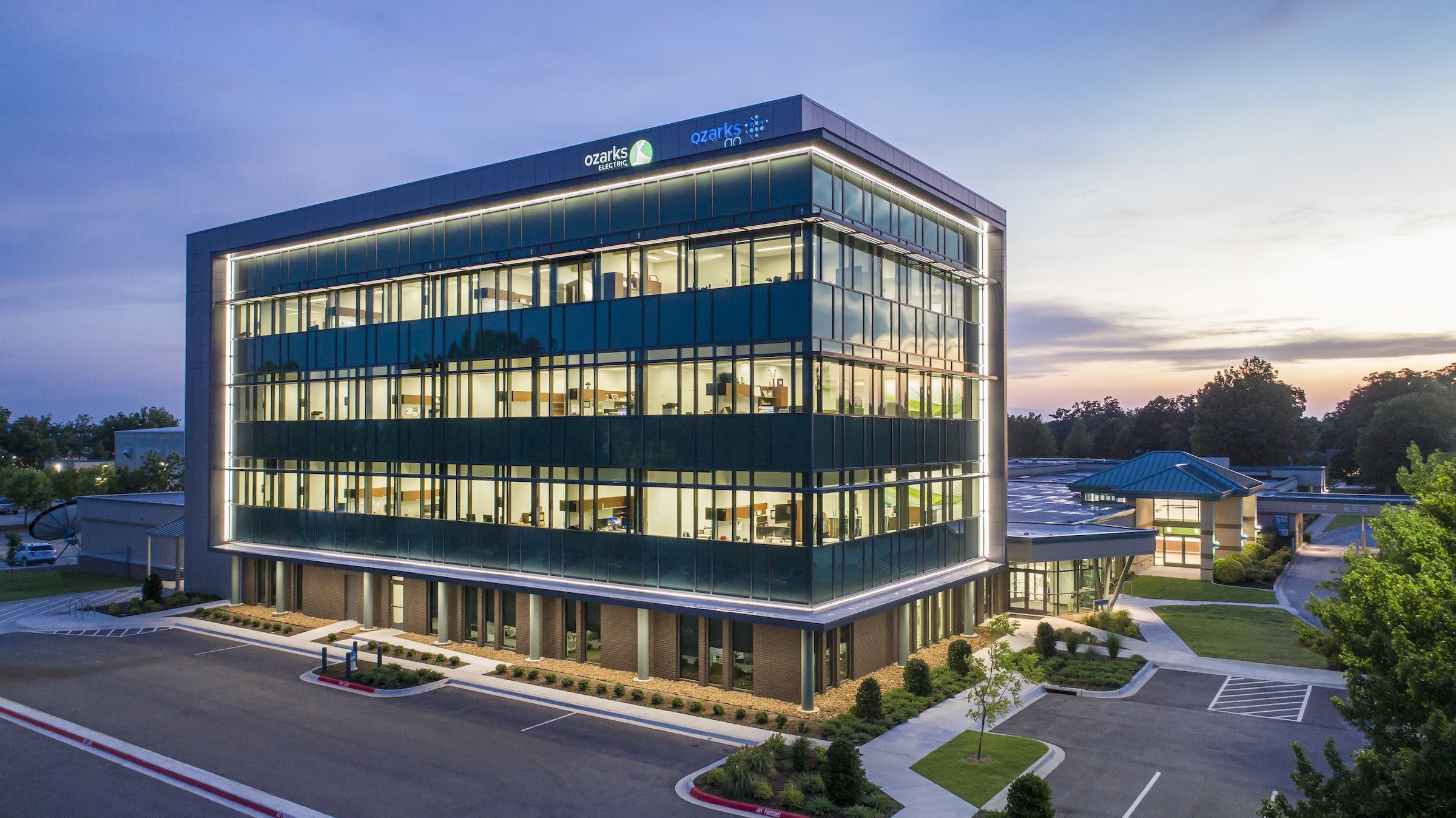 Modern four-story glass office building with illuminated edges, surrounded by a parking lot and trees at dusk.