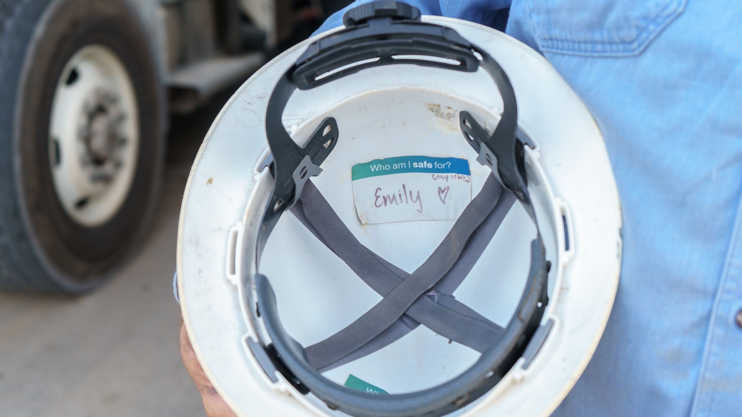A person holds a white hard hat with a name tag inside, reading "Emily" with a heart. A truck tire is visible in the background.