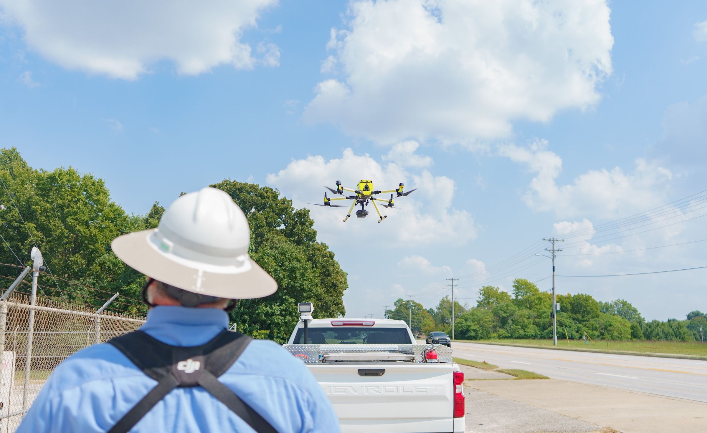 A person in a hard hat operates a drone near a road, with trees and a white truck in the background under a cloudy sky.