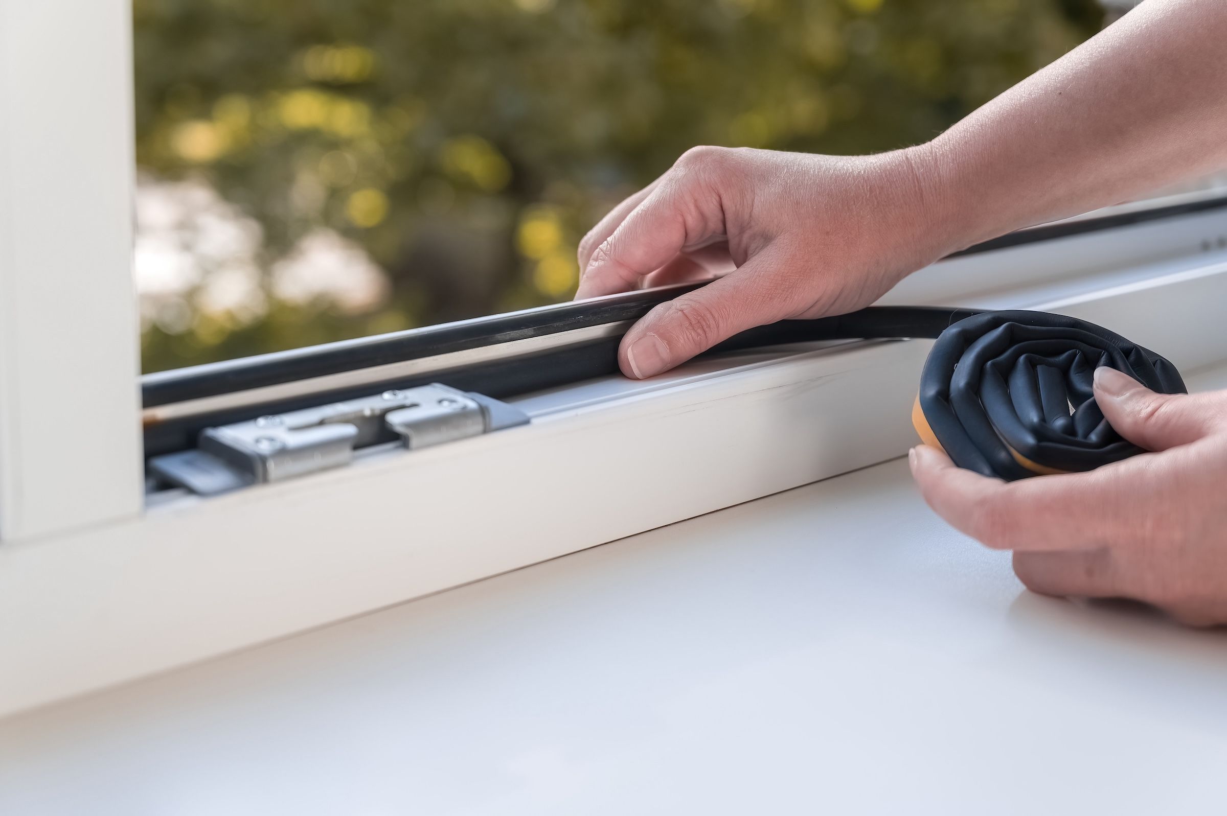 Hands installing weatherstripping on a white window frame, with a coiled strip nearby.
