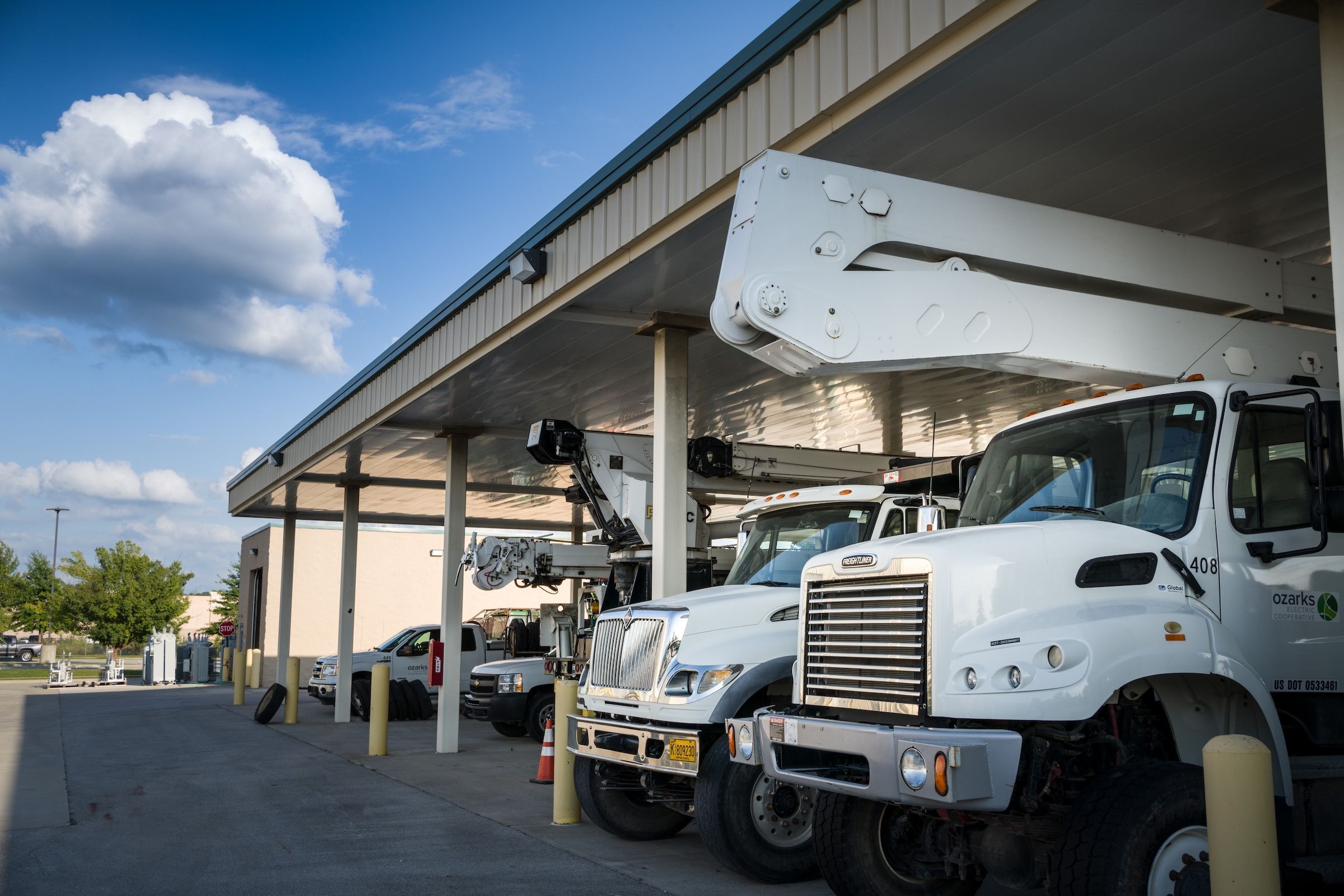 Utility trucks parked under a large canopy at a service facility, with a partly cloudy sky in the background.