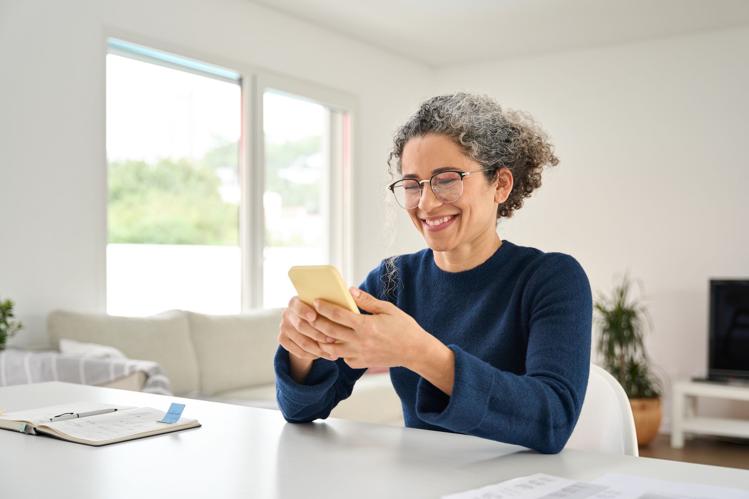 Smiling woman with curly hair and glasses using a smartphone at a table in a bright living room.