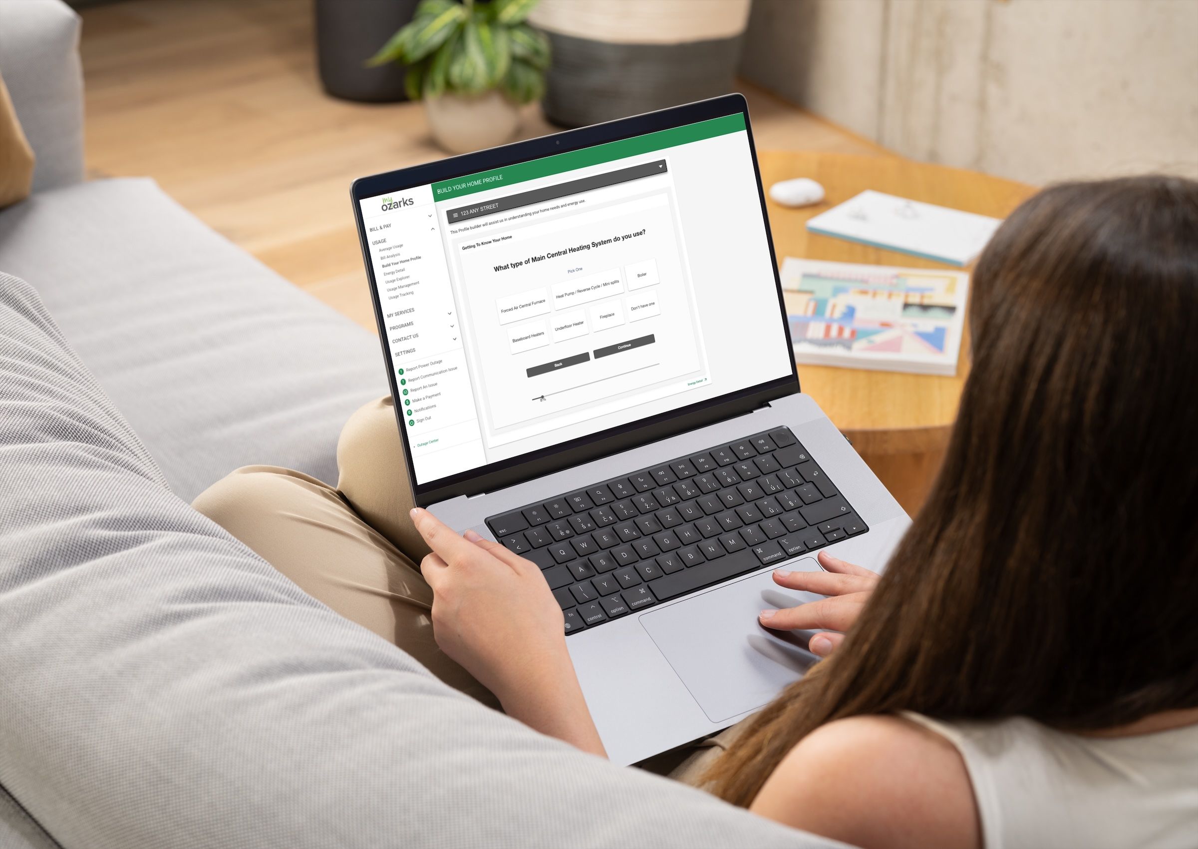 Person sitting on a couch using a laptop displaying a software interface, with a wooden table and a plant in the background.