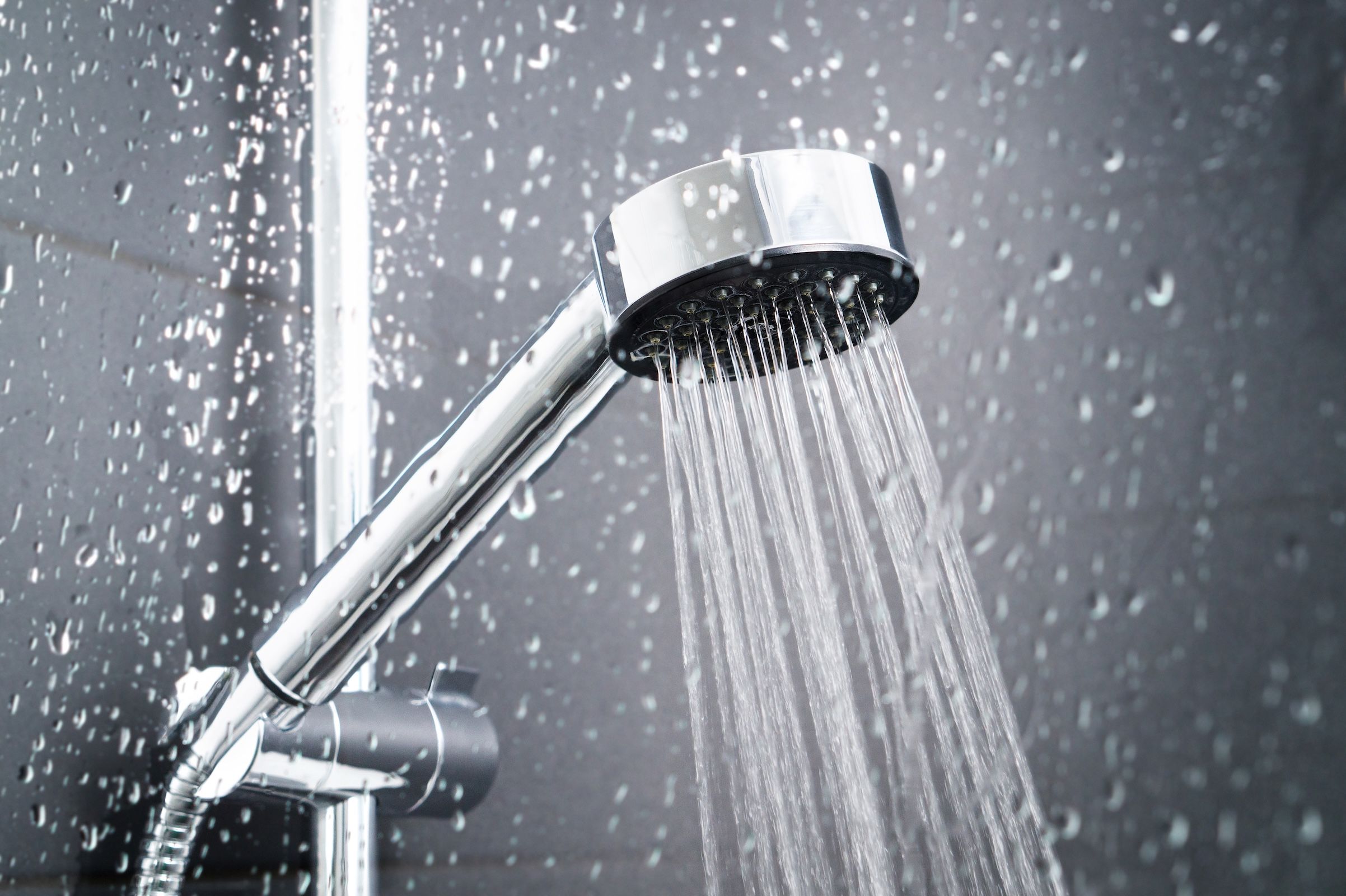 A chrome showerhead spraying water, with droplets visible on the dark tiled wall background.