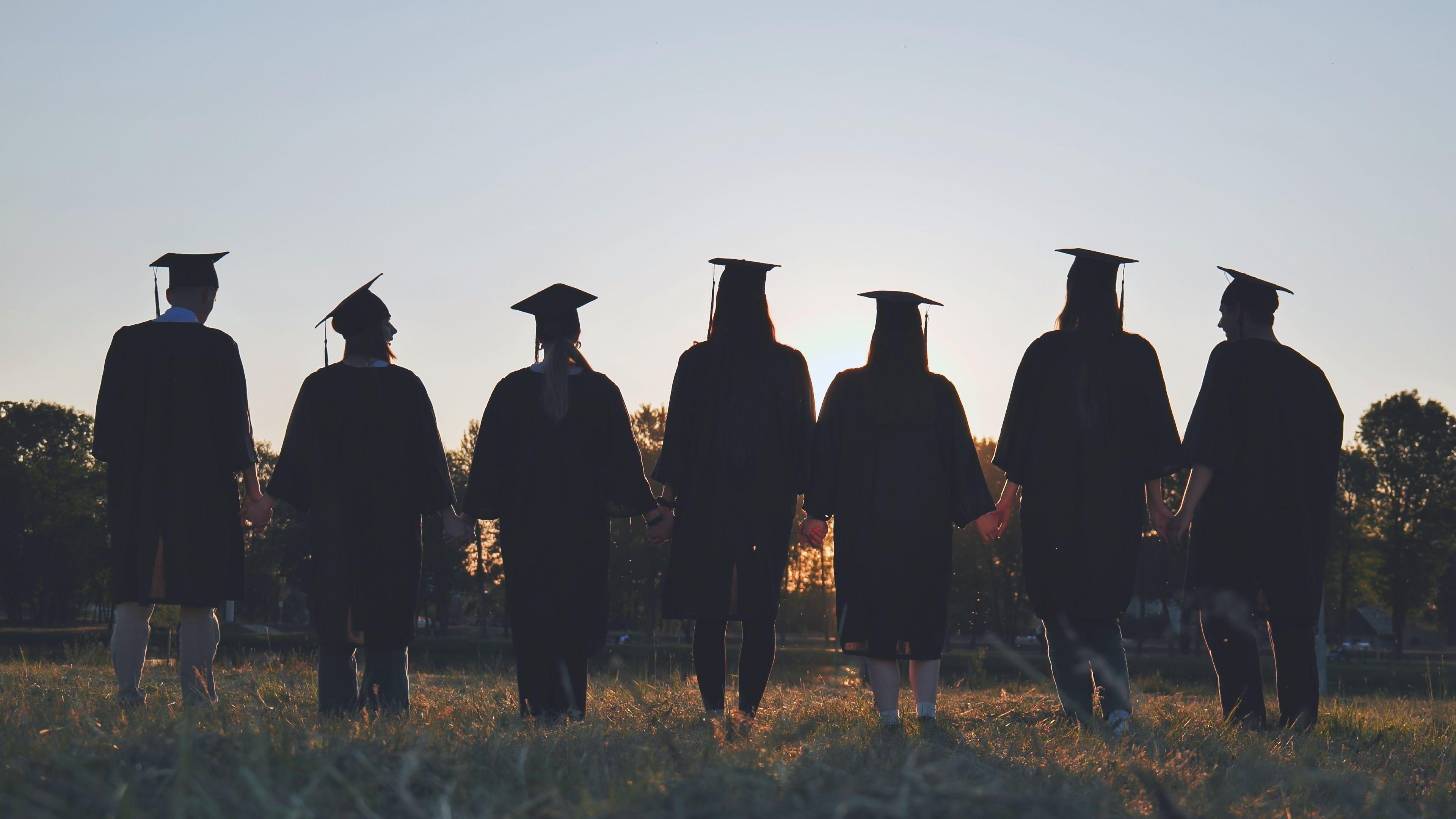 Seven graduates in caps and gowns stand in a grassy field, facing away, with a sunset in the background.