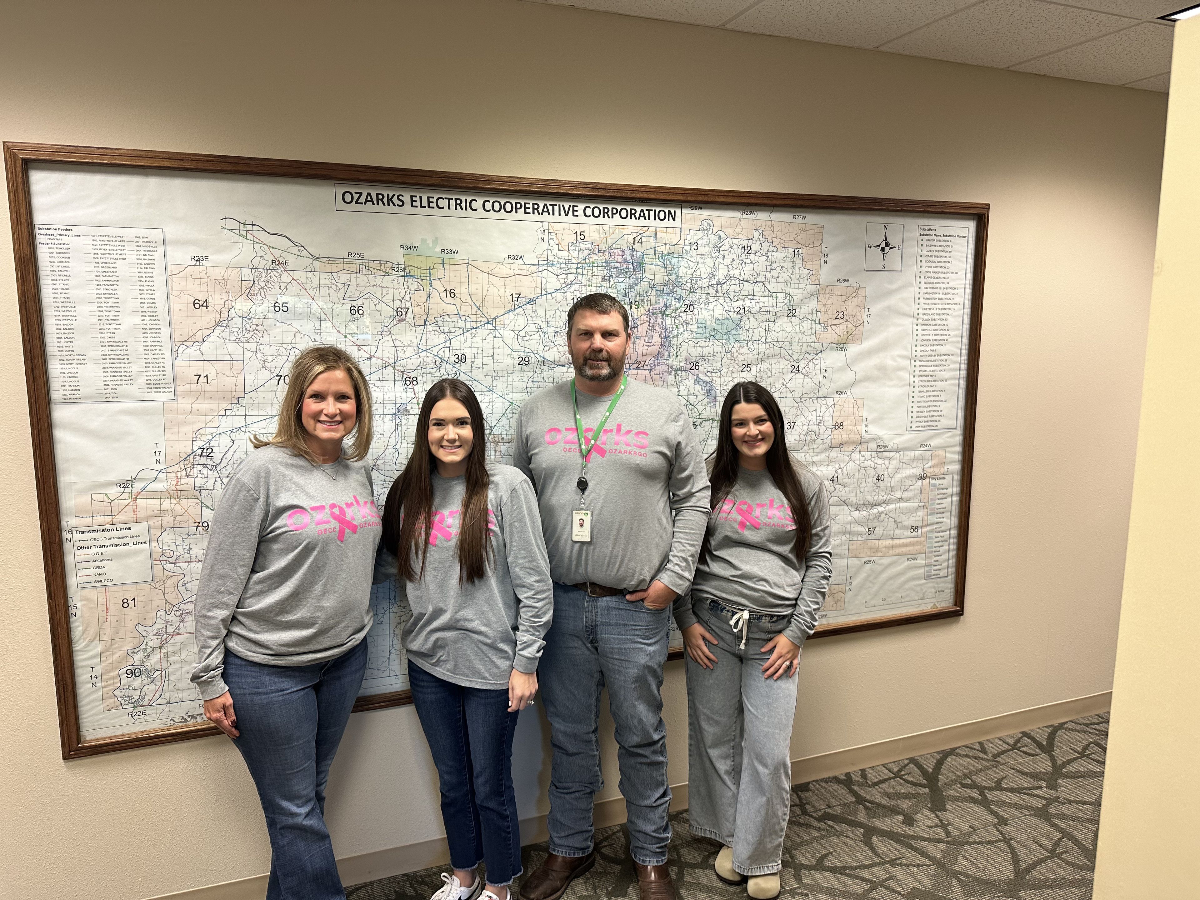 Four people in matching gray shirts stand smiling in front of a large wall map at Ozarks Electric Cooperative Corporation.