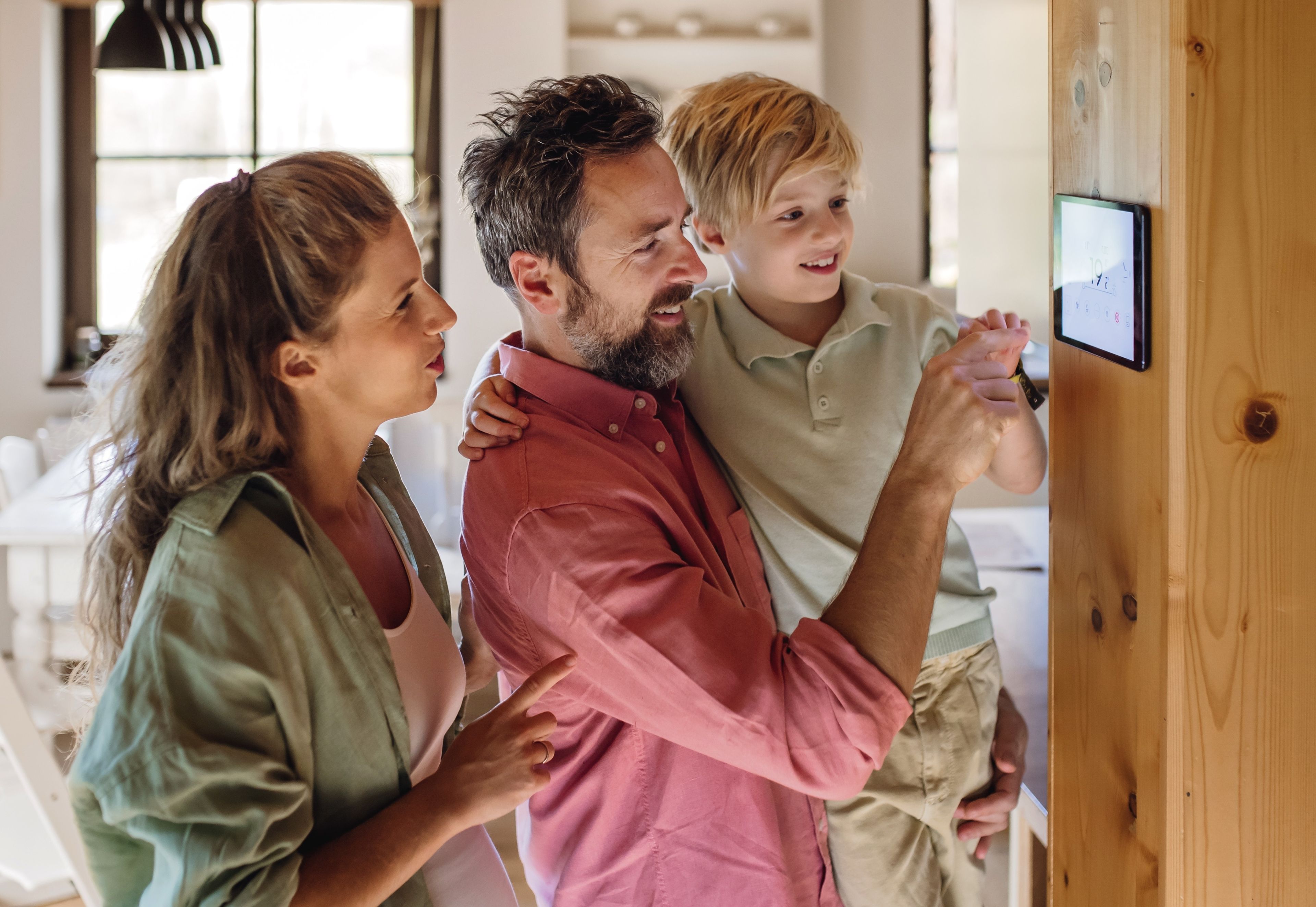 A family of three adjusts a smart home device on the wall in a cozy, well-lit living room. The parents smile as their child interacts with the screen.