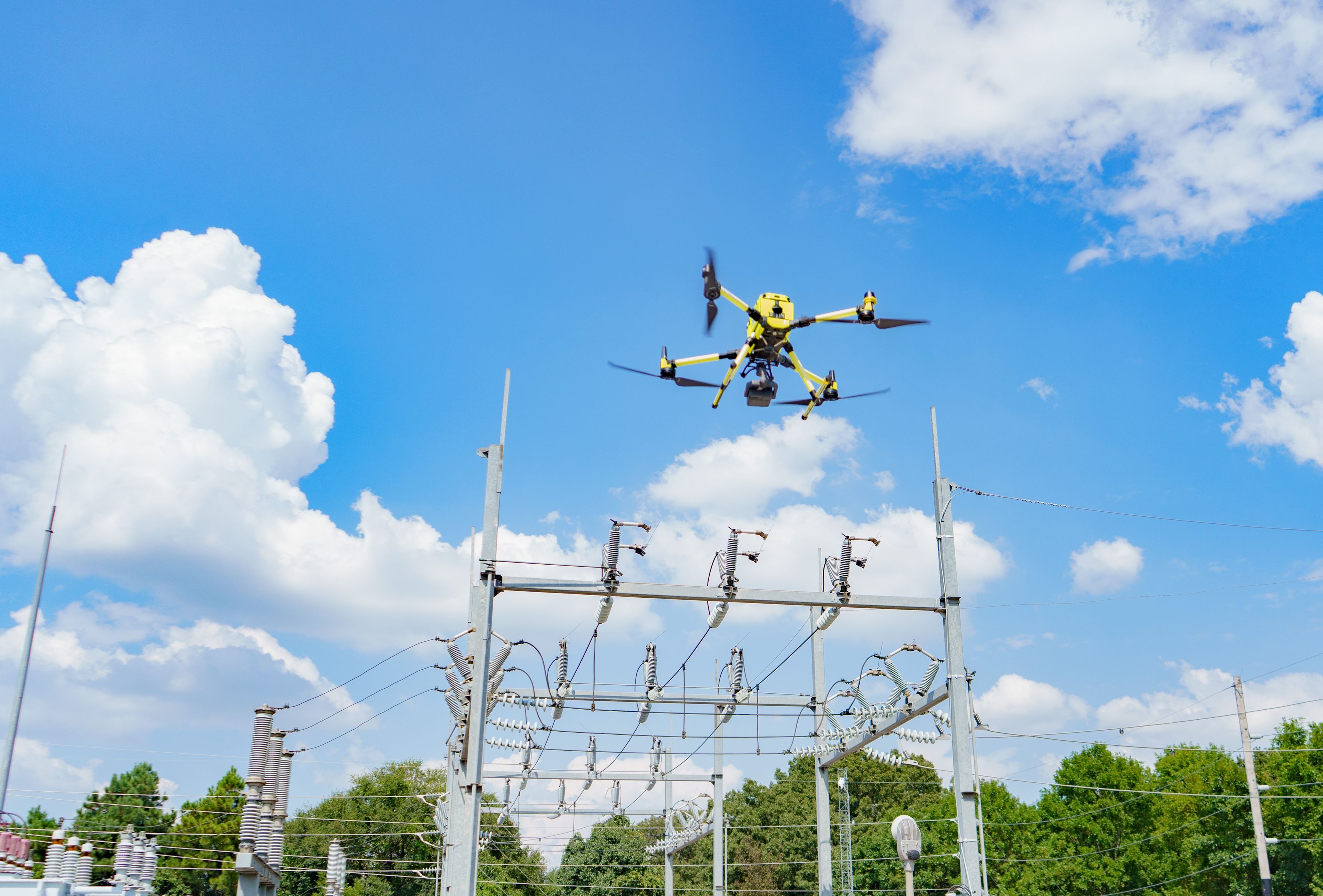 A yellow drone flying over electrical power lines against a blue sky with clouds and trees in the background.