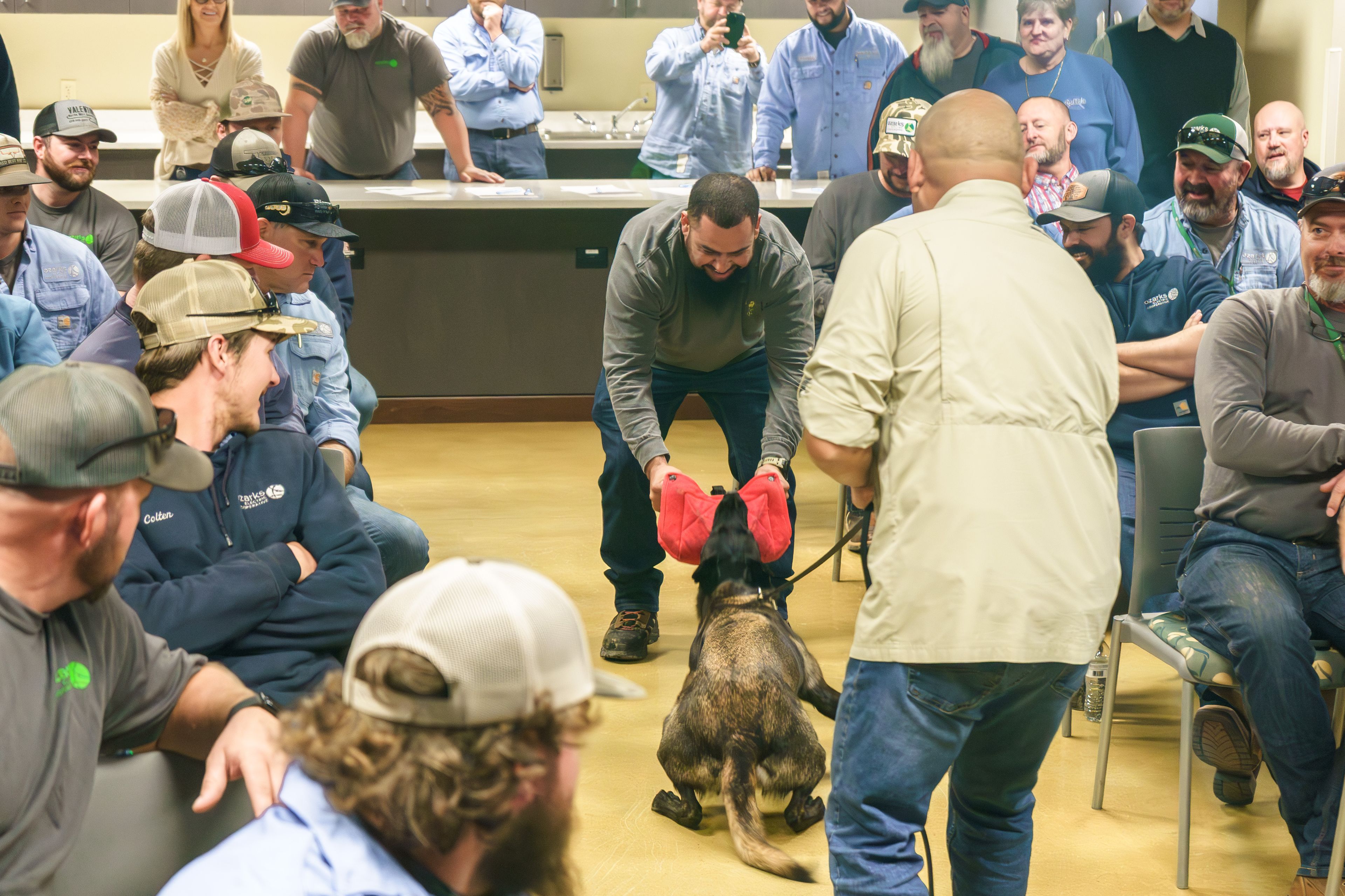 A group of people watch a man playing tug-of-war with a dog using a red toy in a casual indoor setting.