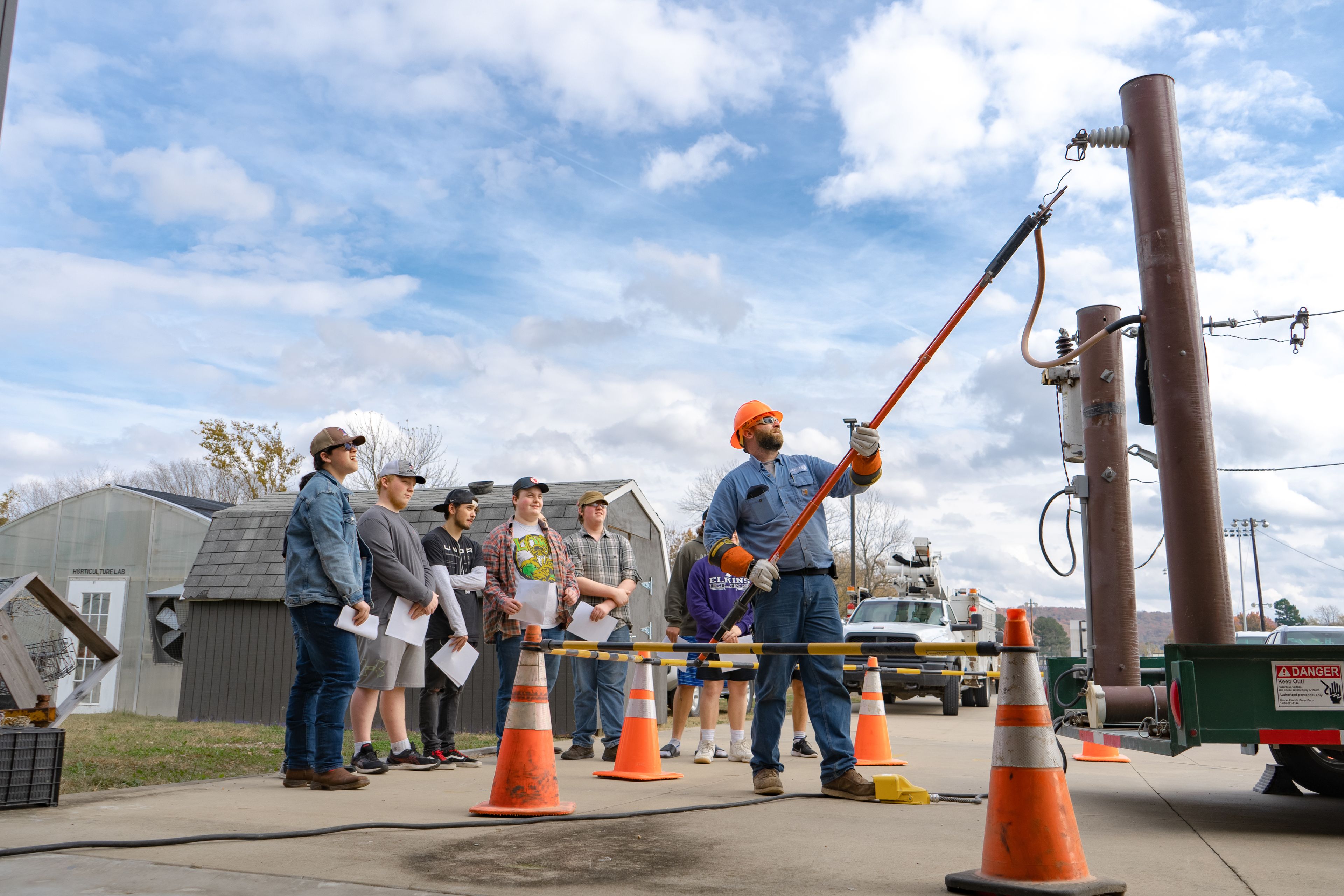A worker demonstrates electrical safety to a group outdoors, using a long tool near power lines, with traffic cones marking the area.