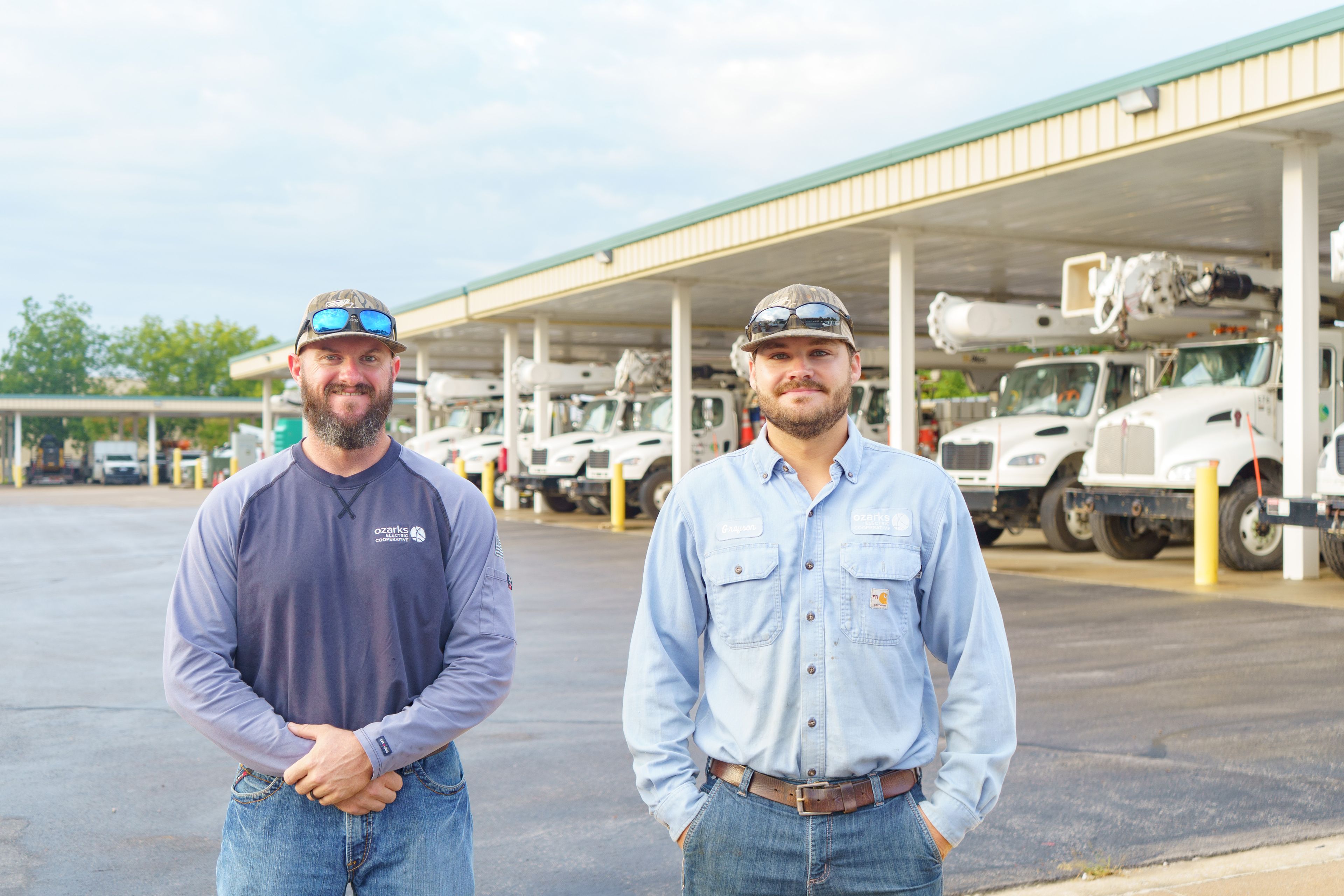 Two men in casual work attire stand in front of a row of utility trucks under a covered area in a parking lot.