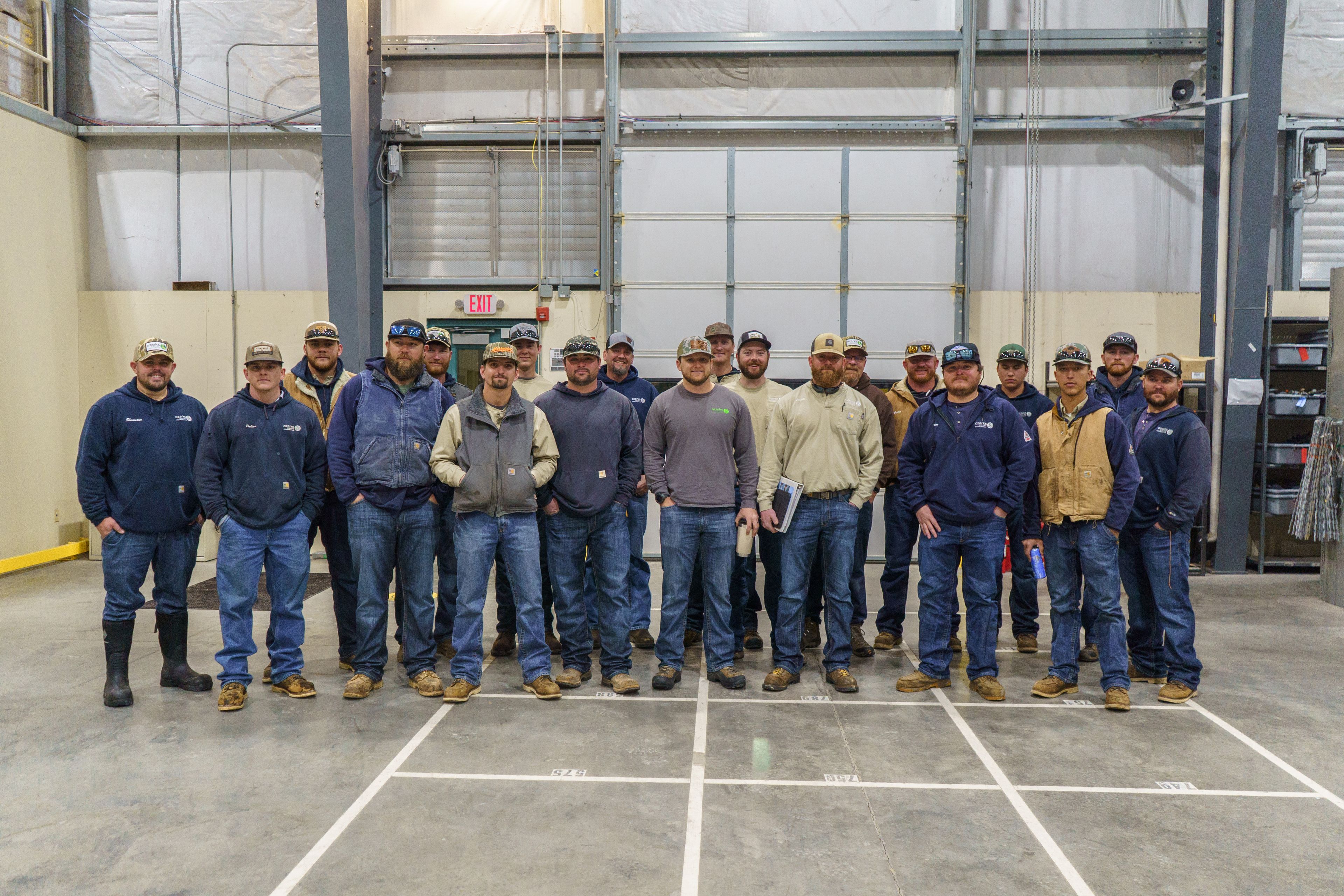 A group of 17 people wearing casual work attire stands in a large industrial space with high ceilings and gray partitions.