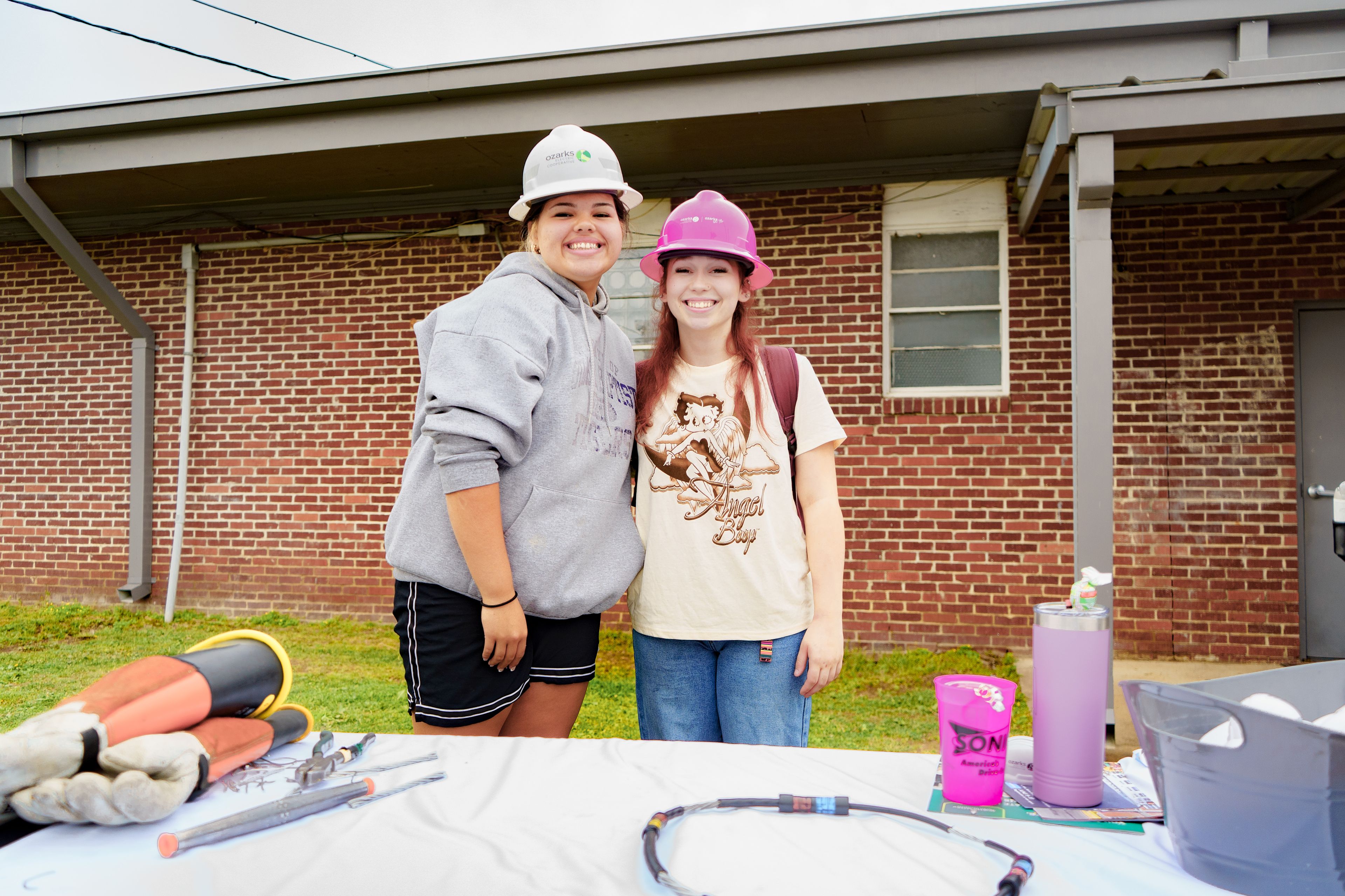 Two people wearing hard hats smile at the camera. They stand in front of a brick building, with tools and drinks on a table nearby.