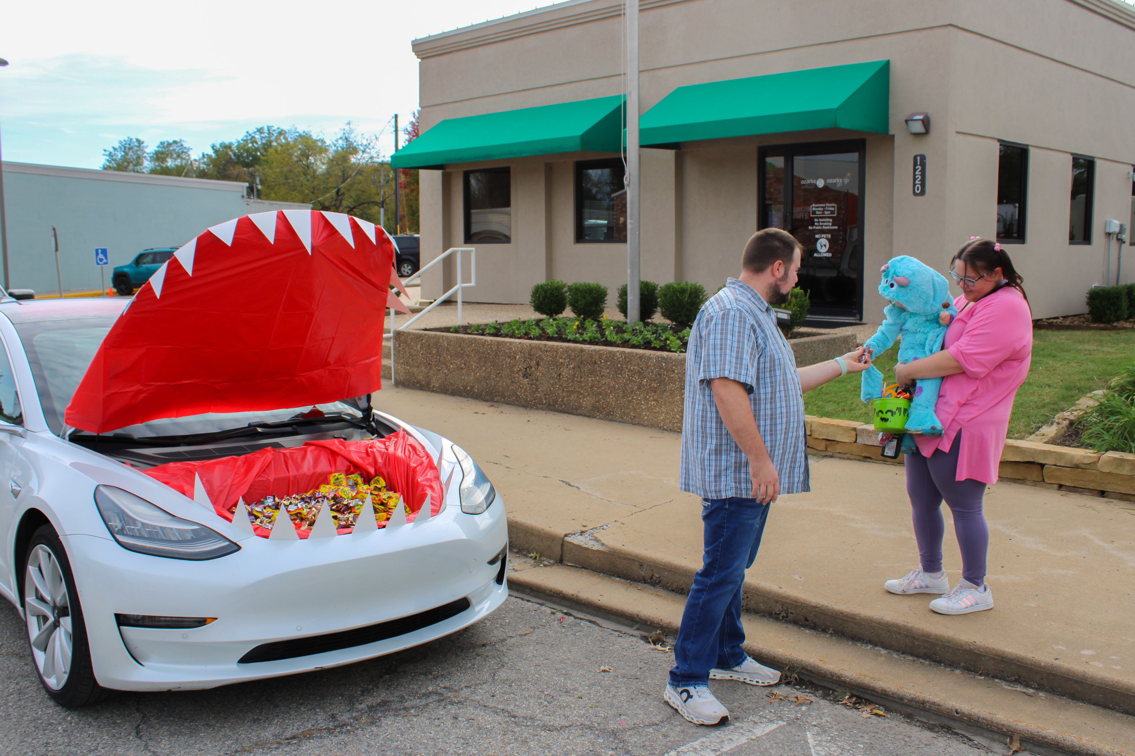 Car trunk decorated as a shark mouth with candy inside; a man hands candy to a woman holding a child in a costume outside a building.