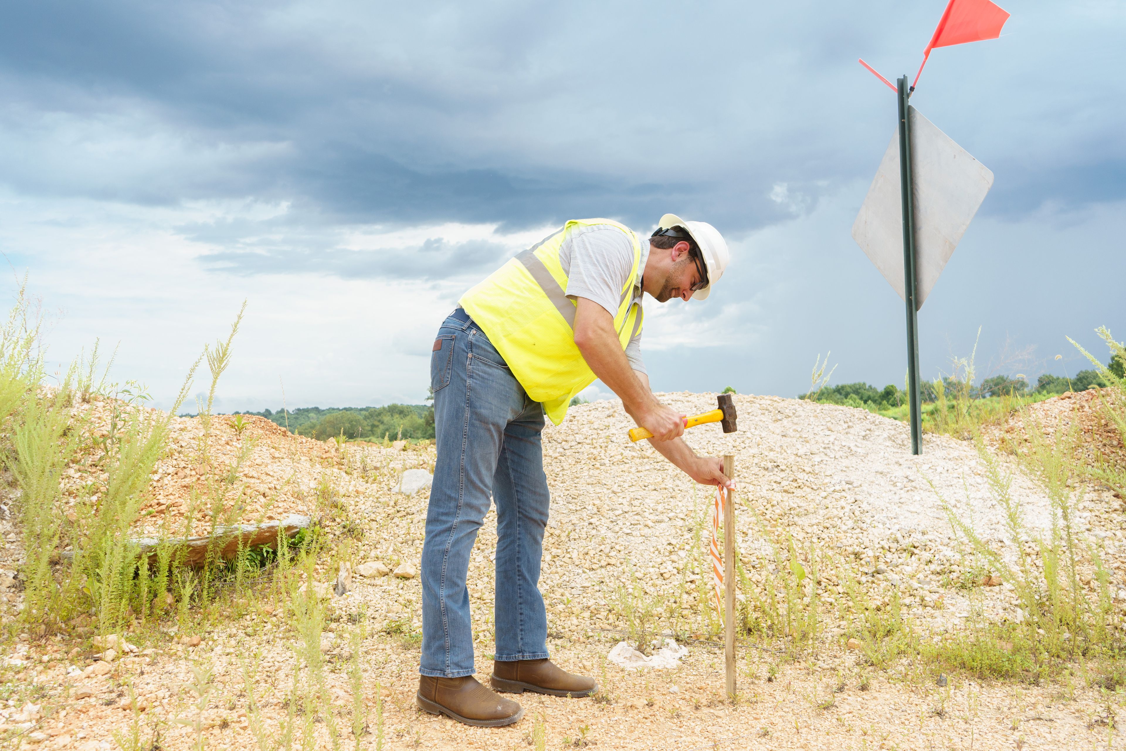 Worker in a yellow vest and hard hat hammers a stake into the ground at a construction site, with a red flag and cloudy sky in the background.