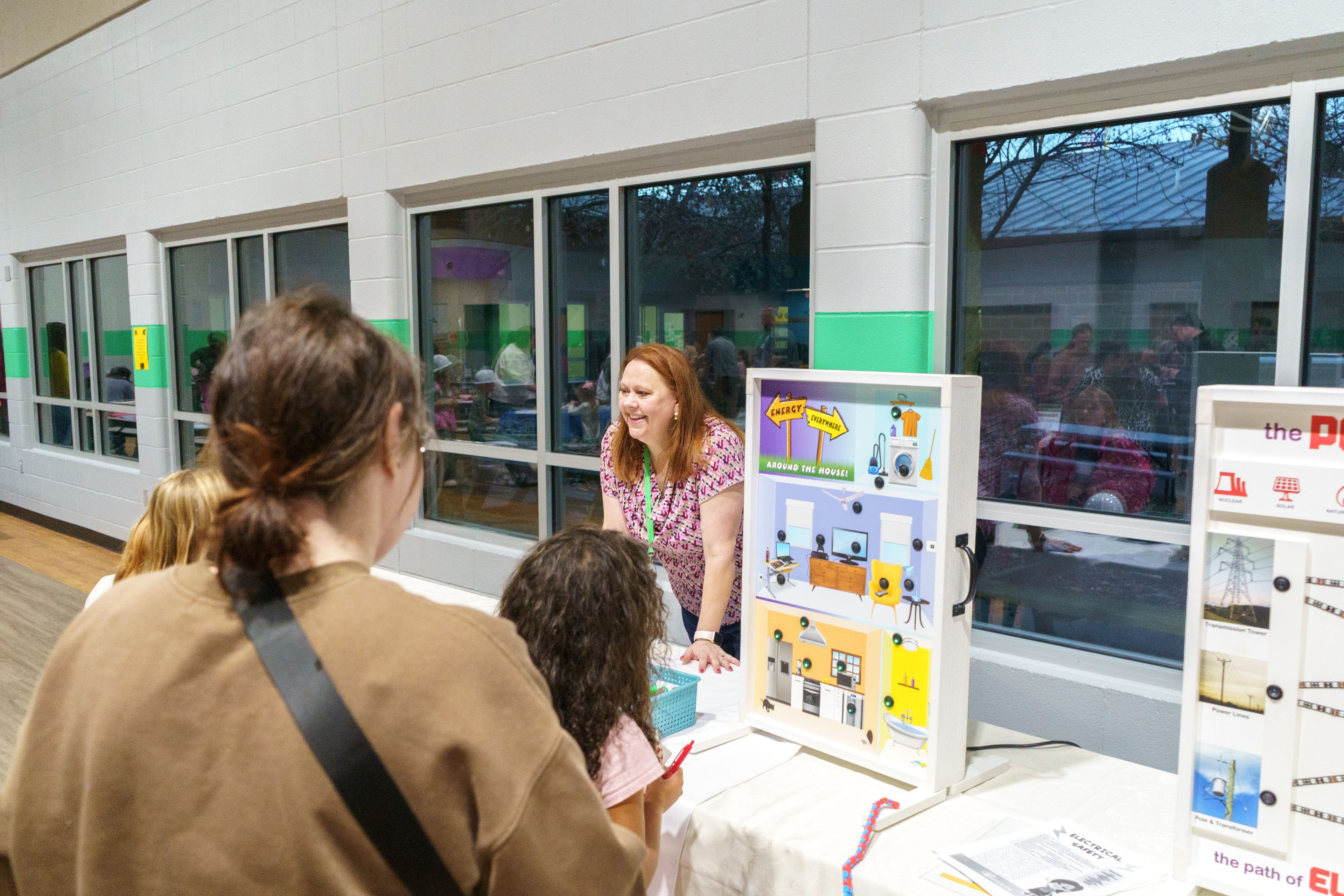 A woman with a green lanyard behind a table smiles at a group of people admiring the displays.