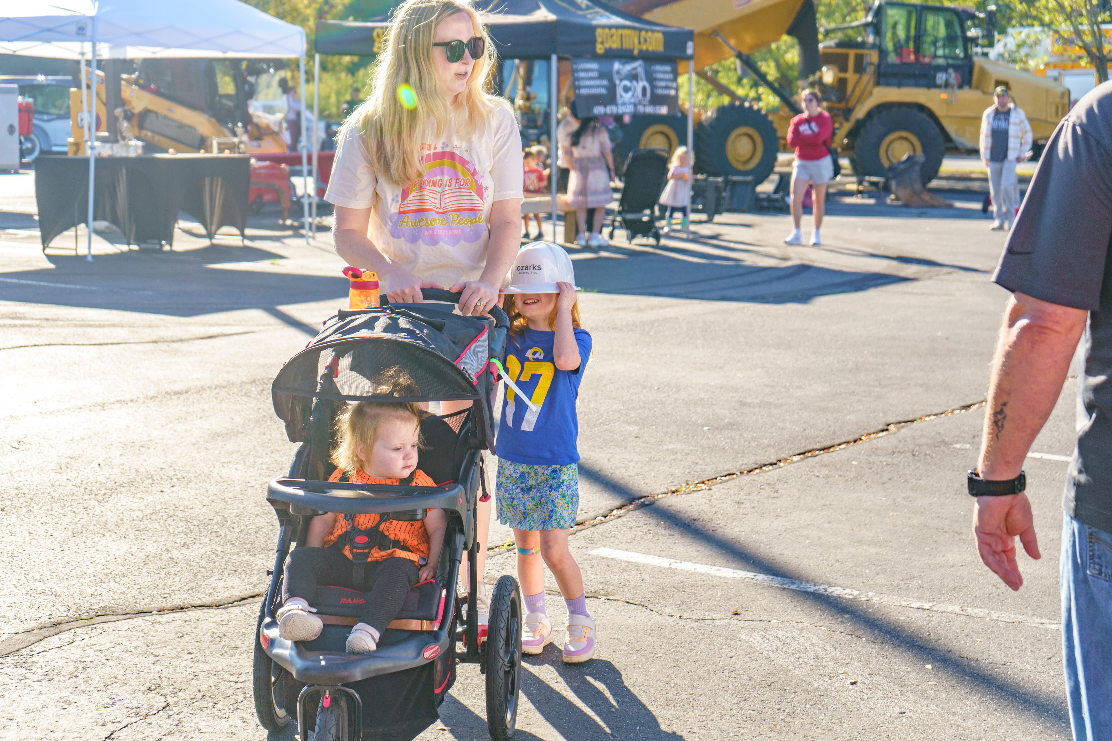 A woman pushes a stroller with a child, while another child walks beside them, wearing a hat. Construction vehicles are visible in the background.