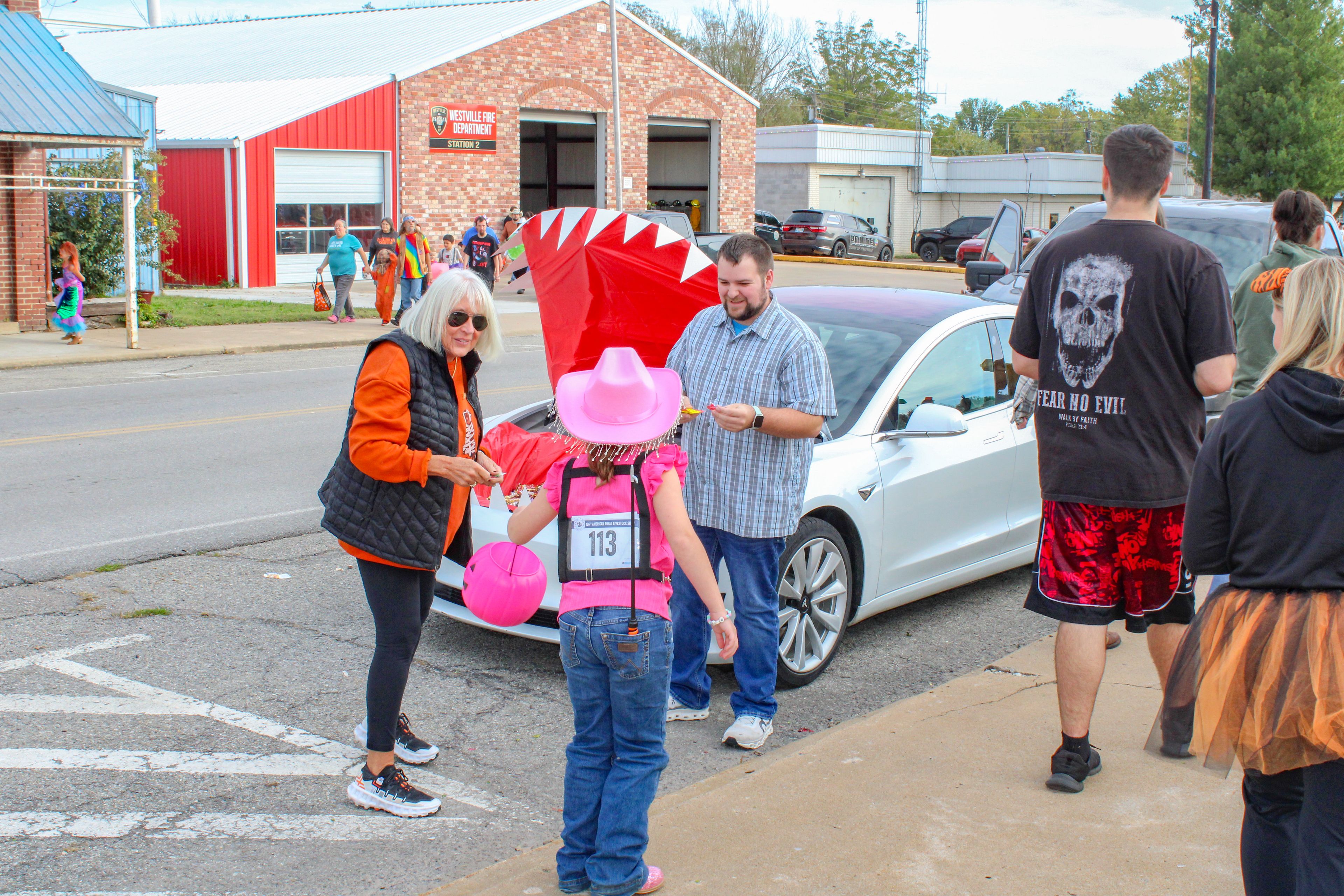 People in costumes gather near a white car on a street. A child in a pink hat and a person with a red headdress stand out.