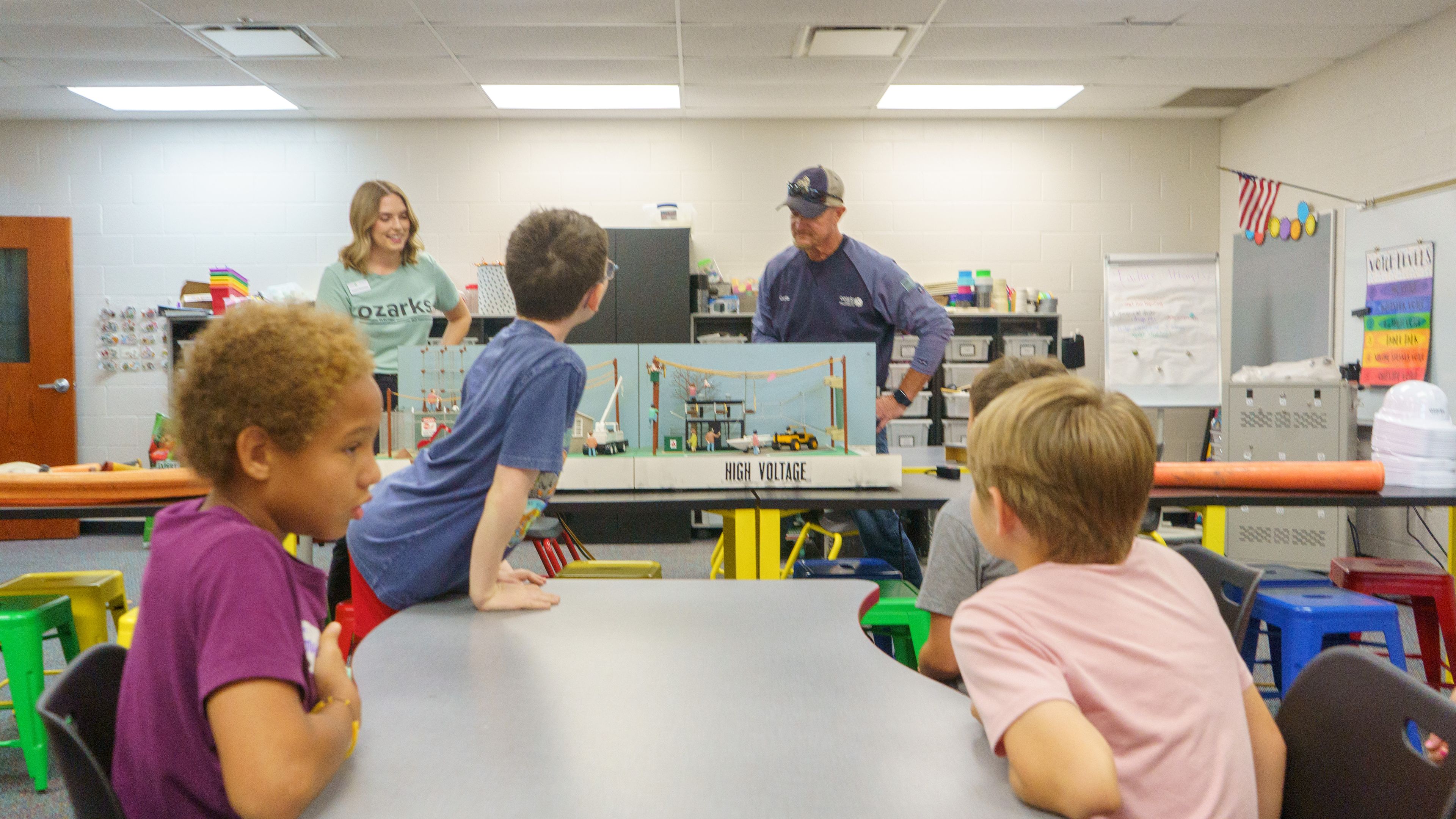 Children in a classroom listen attentively to two adults demonstrating a model construction site, with colorful decorations and furniture around them.