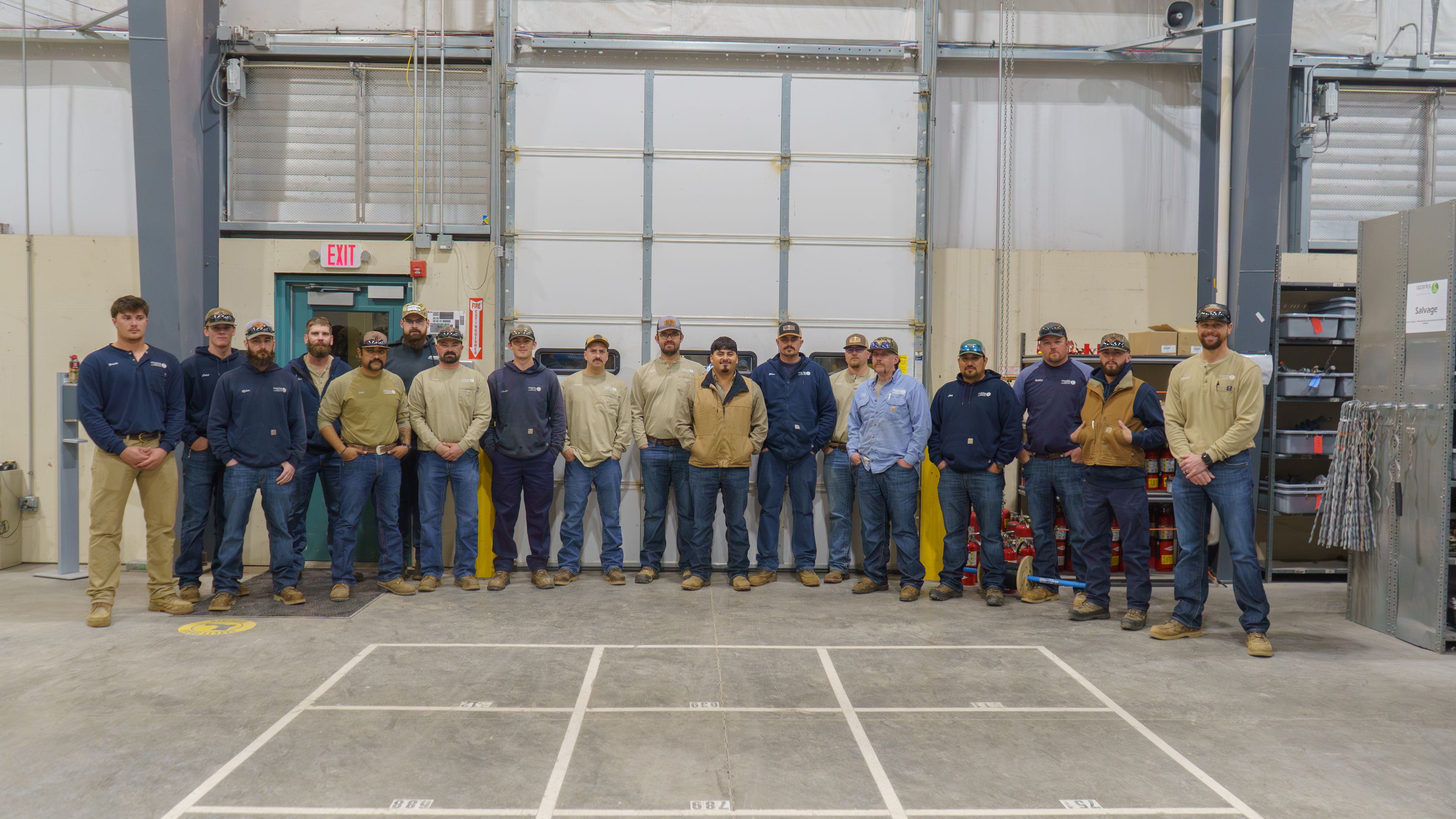 A group of men in work attire stand in a line inside a large industrial space with a garage door and safety equipment in the background.