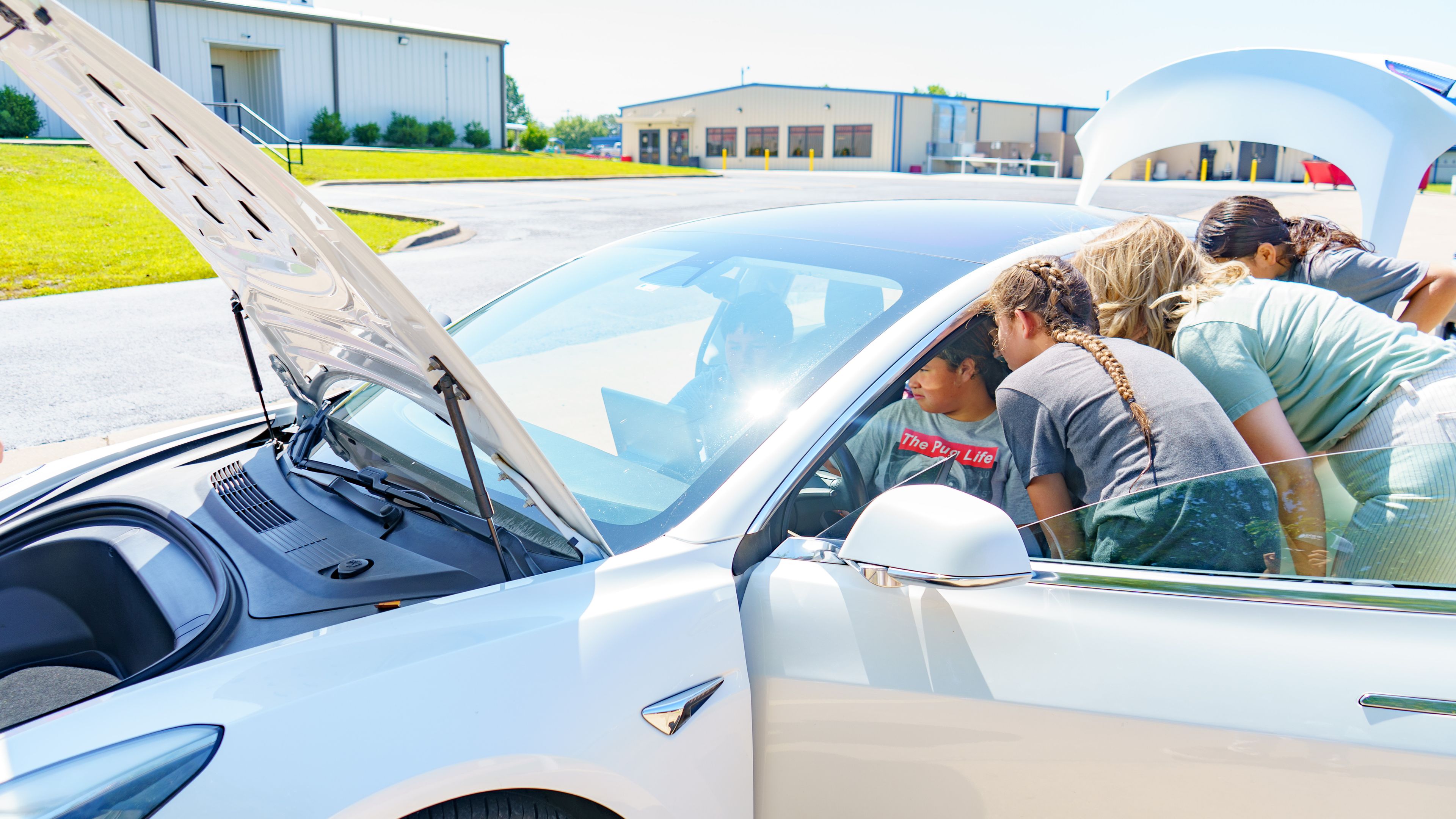 A group of people examine the open front trunk of a white electric vehicle parked outdoors on a sunny day.