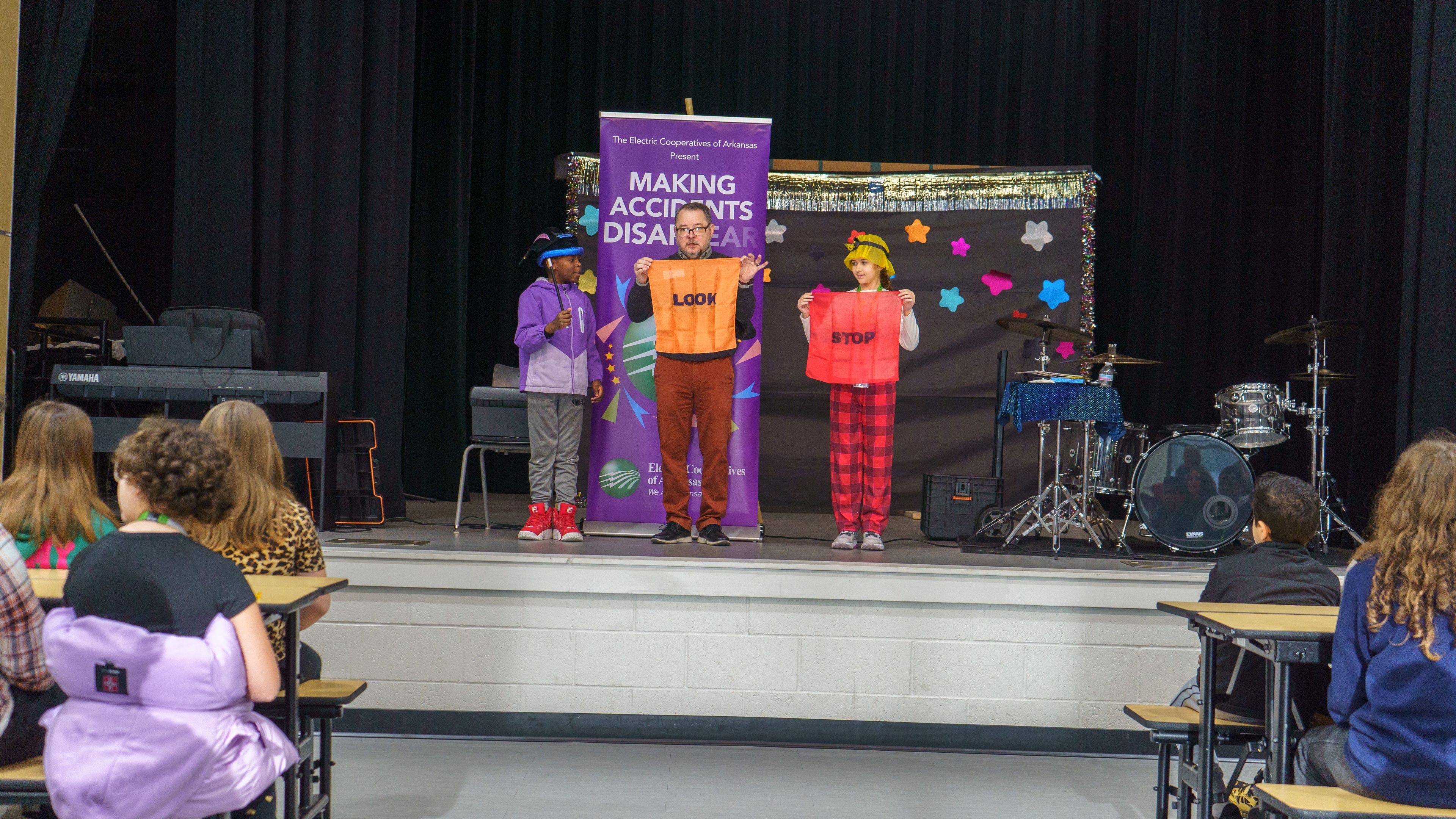 Three performers in colorful costumes stand on stage with a "Making Accidents Disappear" banner, watched by a seated audience.