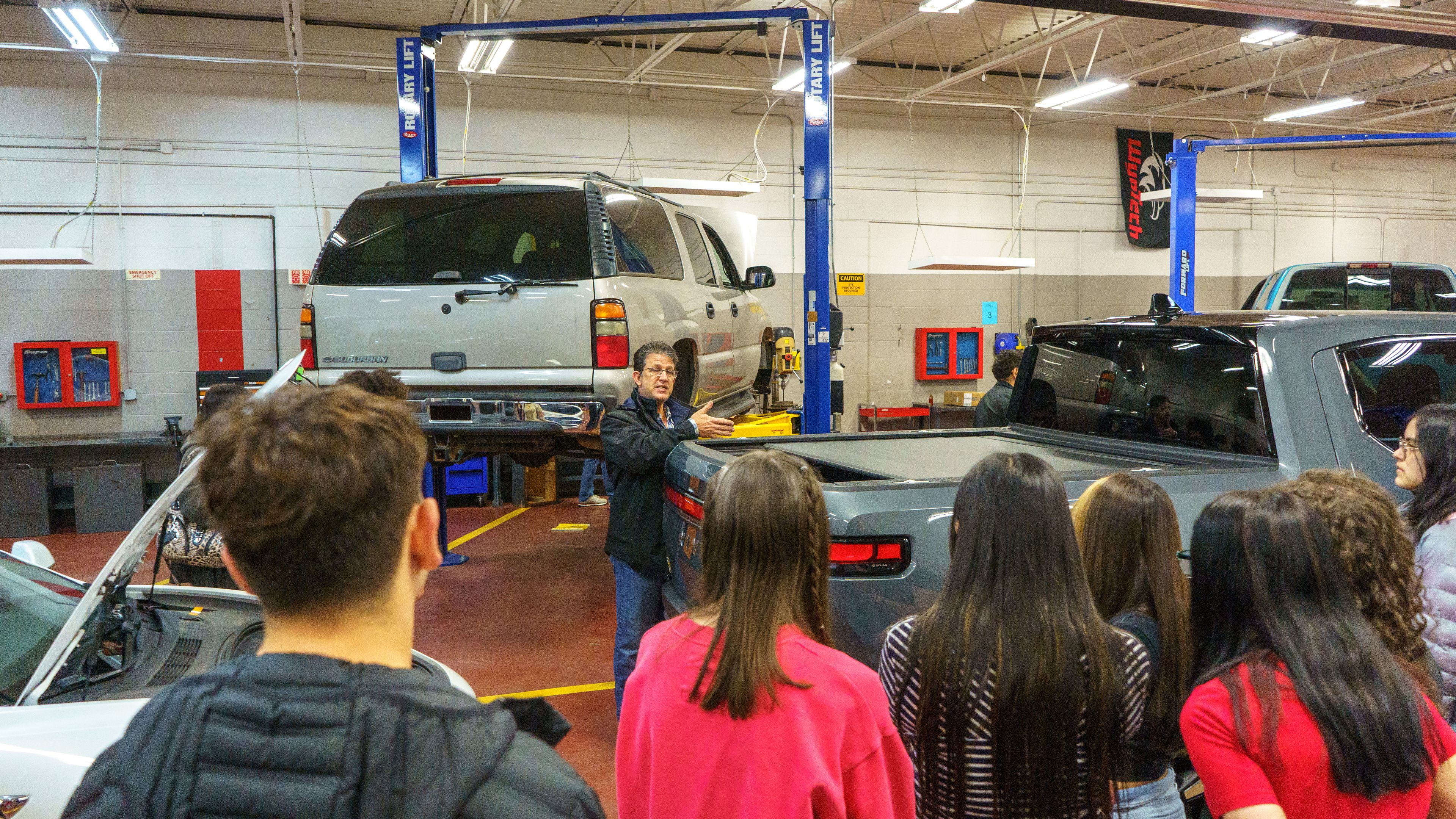 Instructor explaining car maintenance to a group of students in an auto shop with vehicles on lifts.