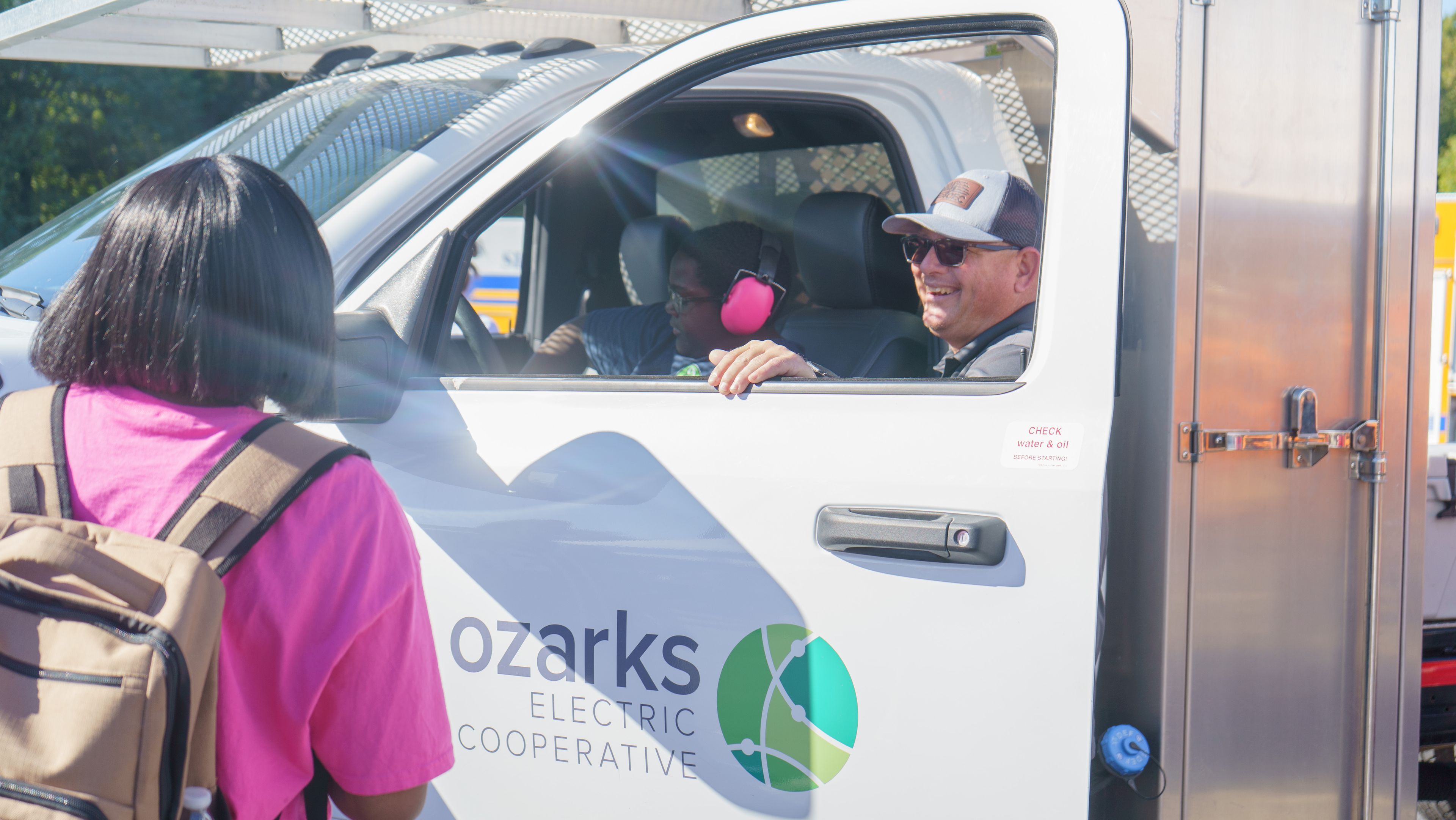 A person in a pink shirt talks to a smiling man in a white Ozarks Electric Cooperative truck on a sunny day.