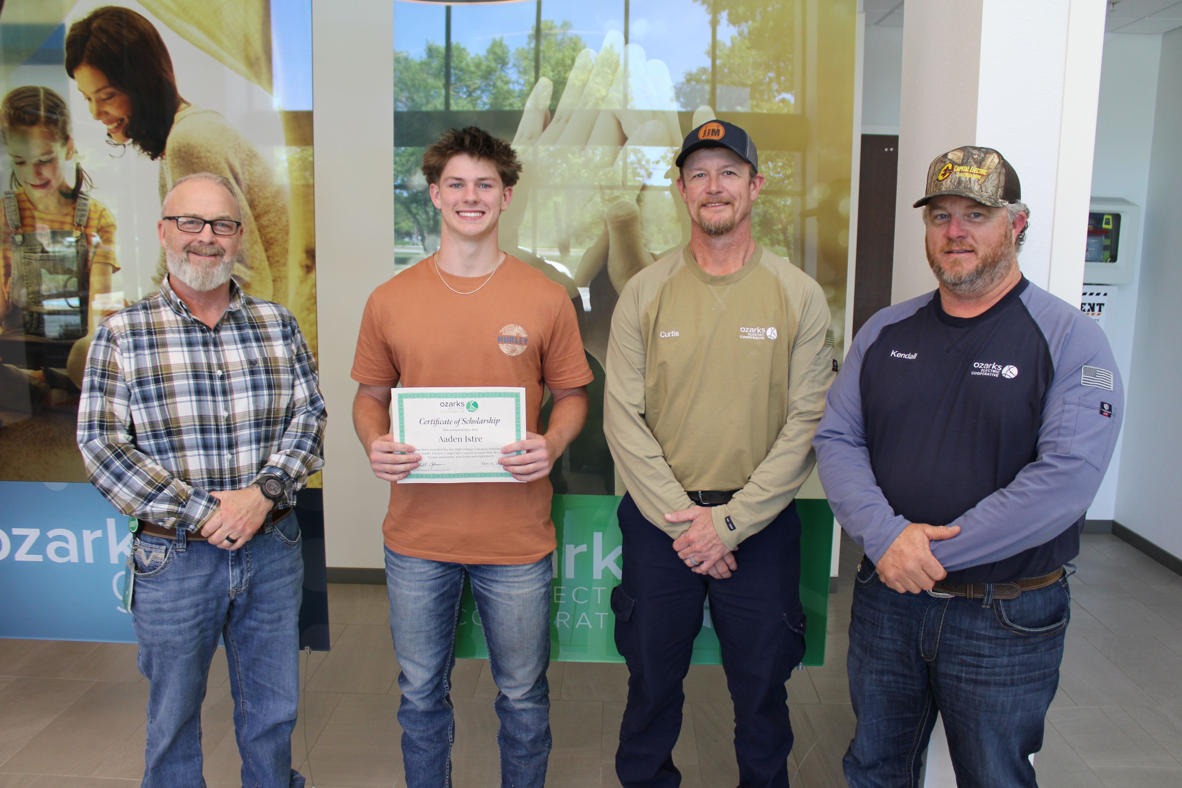 Four men stand indoors; one young man holds a certificate. They smile for the camera in a casual setting.