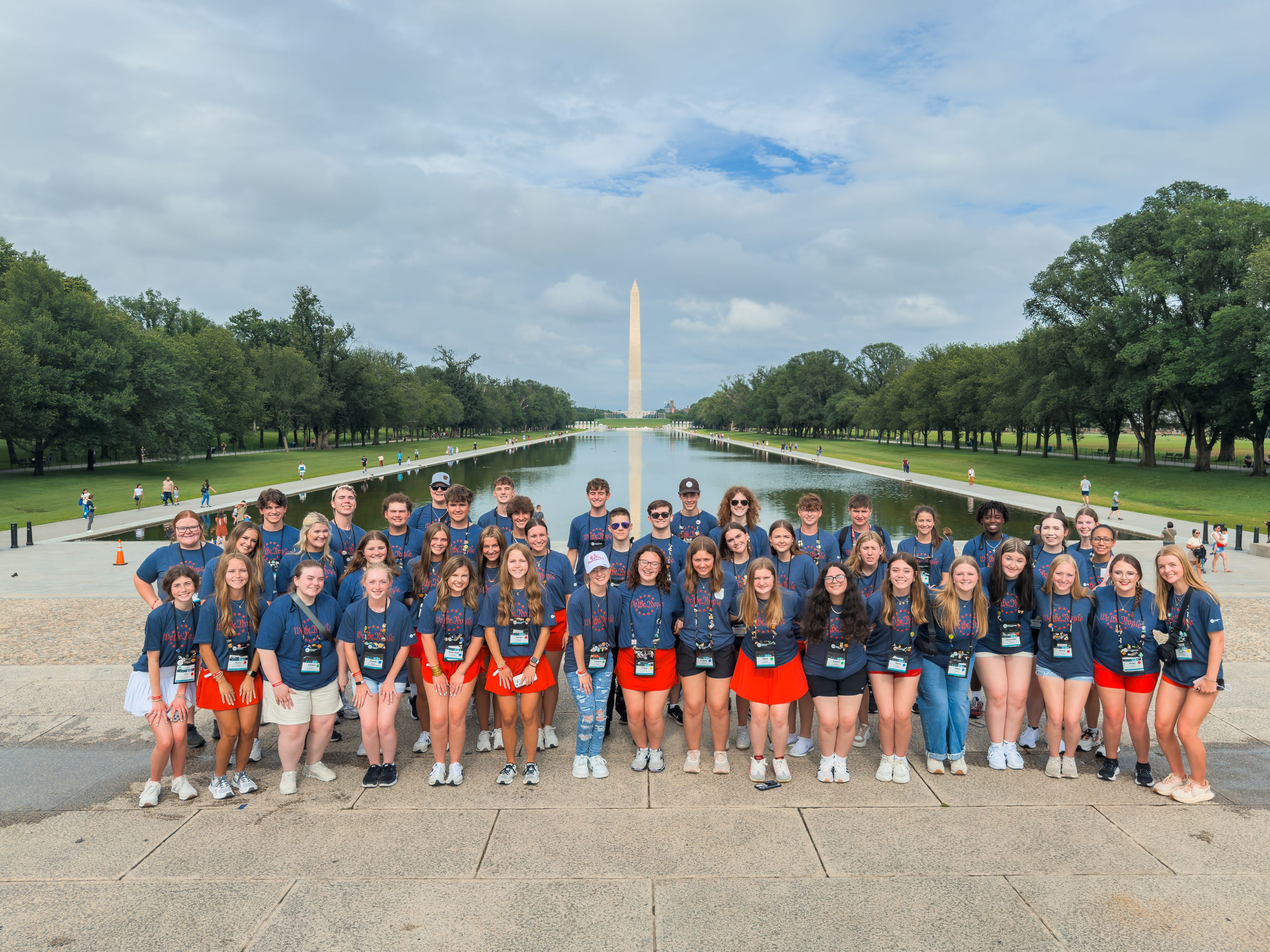 A large group of people in matching blue shirts pose in front of the Washington Monument and Reflecting Pool on a cloudy day.