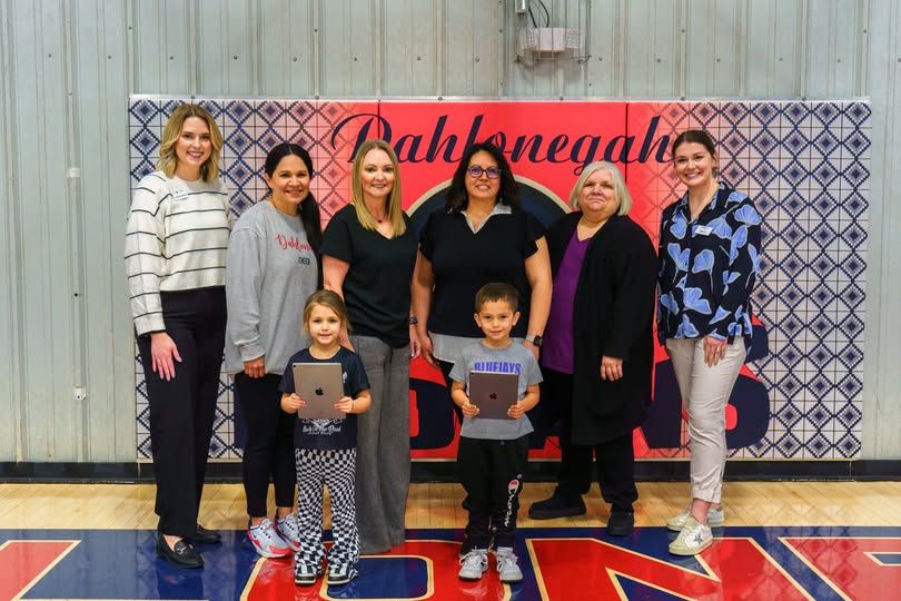 A group of adults and two children holding tablets stand smiling in a gymnasium with a patterned backdrop.