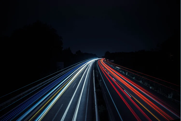 Long-exposure photo of a highway at night, showing white and red light trails from vehicles, creating dynamic lines against a dark sky.