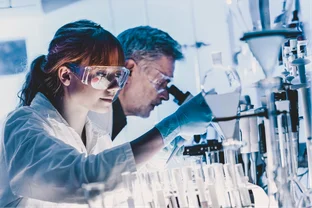 Two scientists in a lab, wearing goggles and gloves, focus on experiments with glassware and microscopes.