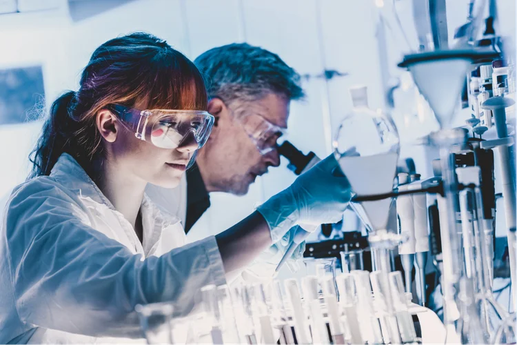 Two scientists in a lab, wearing goggles and gloves, focus on experiments with glassware and microscopes.