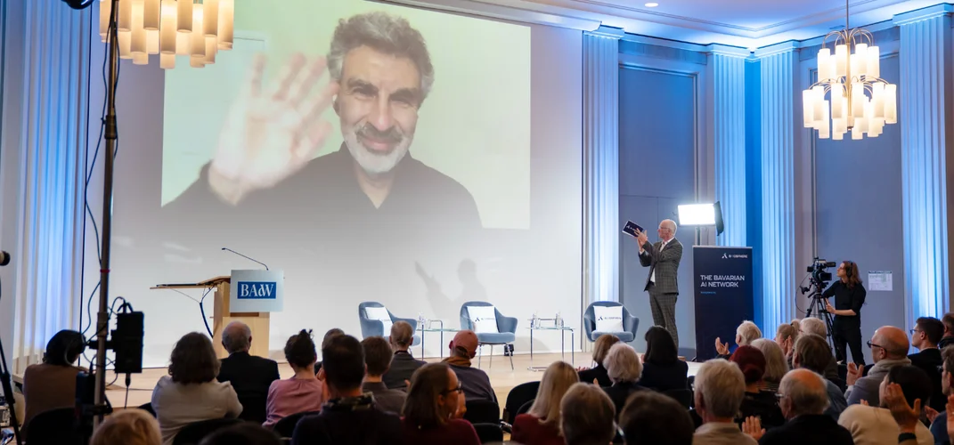 A large crowd watches a man waving on a big screen during an indoor event. A speaker stands near the screen in a well-lit room with chandeliers.