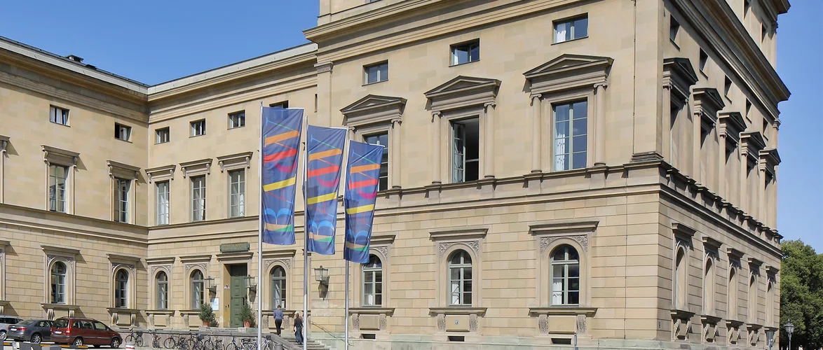 Historisches Steingebäude mit hohen Fenstern und kunstvollen Details, davor zwei bunte Flaggen und links ein geparktes Auto.