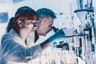 Zwei Wissenschaftler in einem Labor, die Schutzbrillen und Schutzkittel tragen und Experimente mit Reagenzgläsern und einem Mikroskop durchführen.