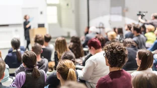A group of people seated in a lecture hall, attentively listening to a speaker at the front, with a camera operator recording the event.