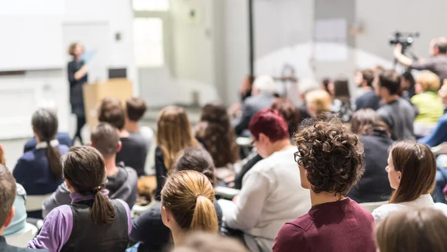 A group of people seated in a lecture hall, attentively listening to a speaker at the front, with a camera operator recording the event.