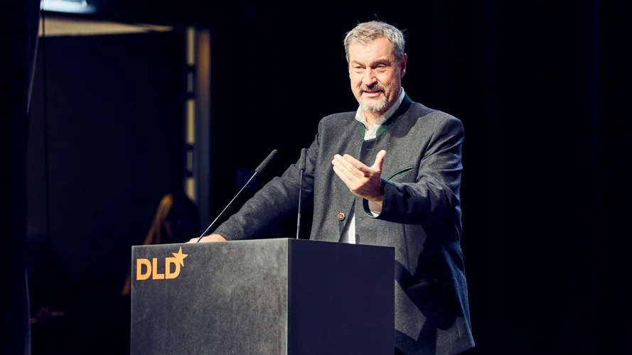 Man speaking at a podium with "DLD" logo, gesturing with one hand. Dark background with stage lighting.
