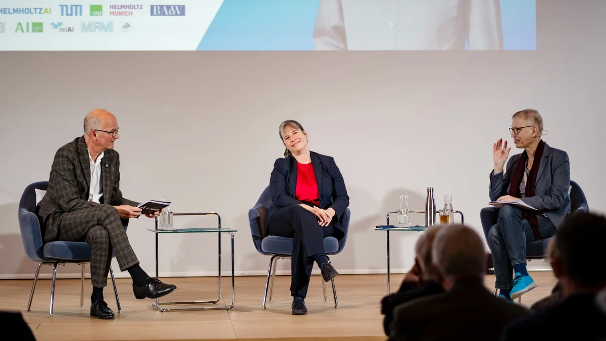 Drei Personen sitzen auf der Bühne in einer Podiumsdiskussion, wobei eine Person spricht und die anderen aufmerksam zuhören.