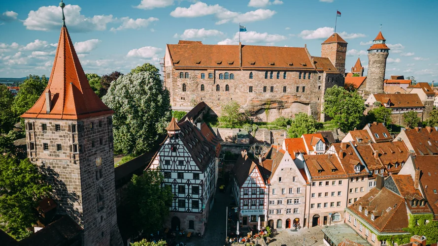 Aerial view of Nuremberg Castle with red-roofed medieval buildings, surrounded by lush greenery under a blue sky with scattered clouds.