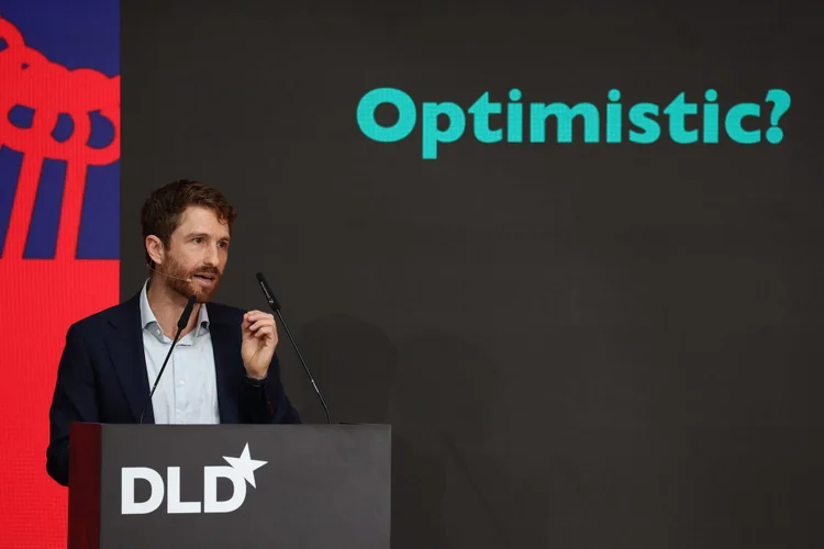 Man speaking at a podium with "DLD" logo, gesturing with his hand. The word "Optimistic?" is displayed on the screen behind him.