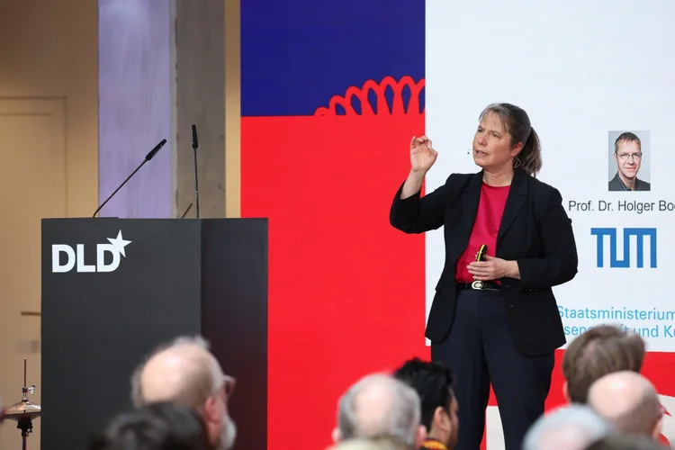 A woman in a suit speaks at a podium labeled "DLD," with a presentation screen behind her displaying text and graphics.