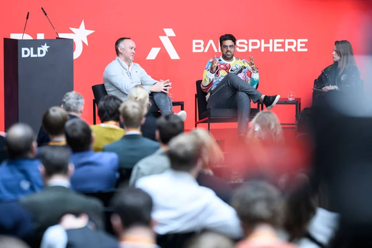 Panel discussion at a conference with three speakers on stage, audience in foreground, and a red backdrop displaying "DLD" and "BACKSPHERE" logos.