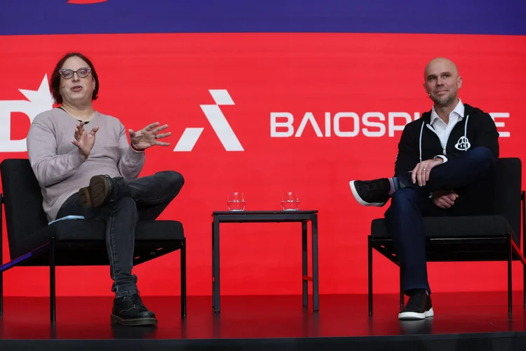 Two individuals seated on stage with microphones, engaging in a discussion against a red backdrop with logos.