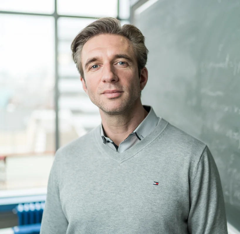 Prof. Kokutta mit kurzen Haaren und einem hellgrauen Pullover steht vor einer Tafel mit mathematischen Gleichungen in einem hellen Raum.