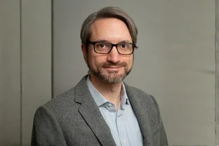 Aaron Johnson mit Brille, kurzen Haaren und Bart, bekleidet mit einem grauen Blazer und einem hellblauen Hemd, steht vor einem einfarbigen Hintergrund.